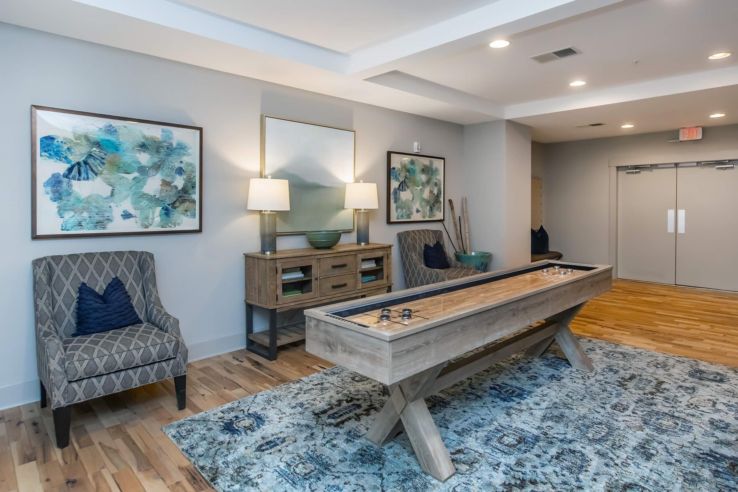 A modern interior scene featuring two patterned chairs next to a wooden console table with decorative items. A shuffleboard table is positioned centrally on a decorative rug, and two framed artworks adorn the wall behind. Soft lighting from table lamps creates a welcoming atmosphere.
