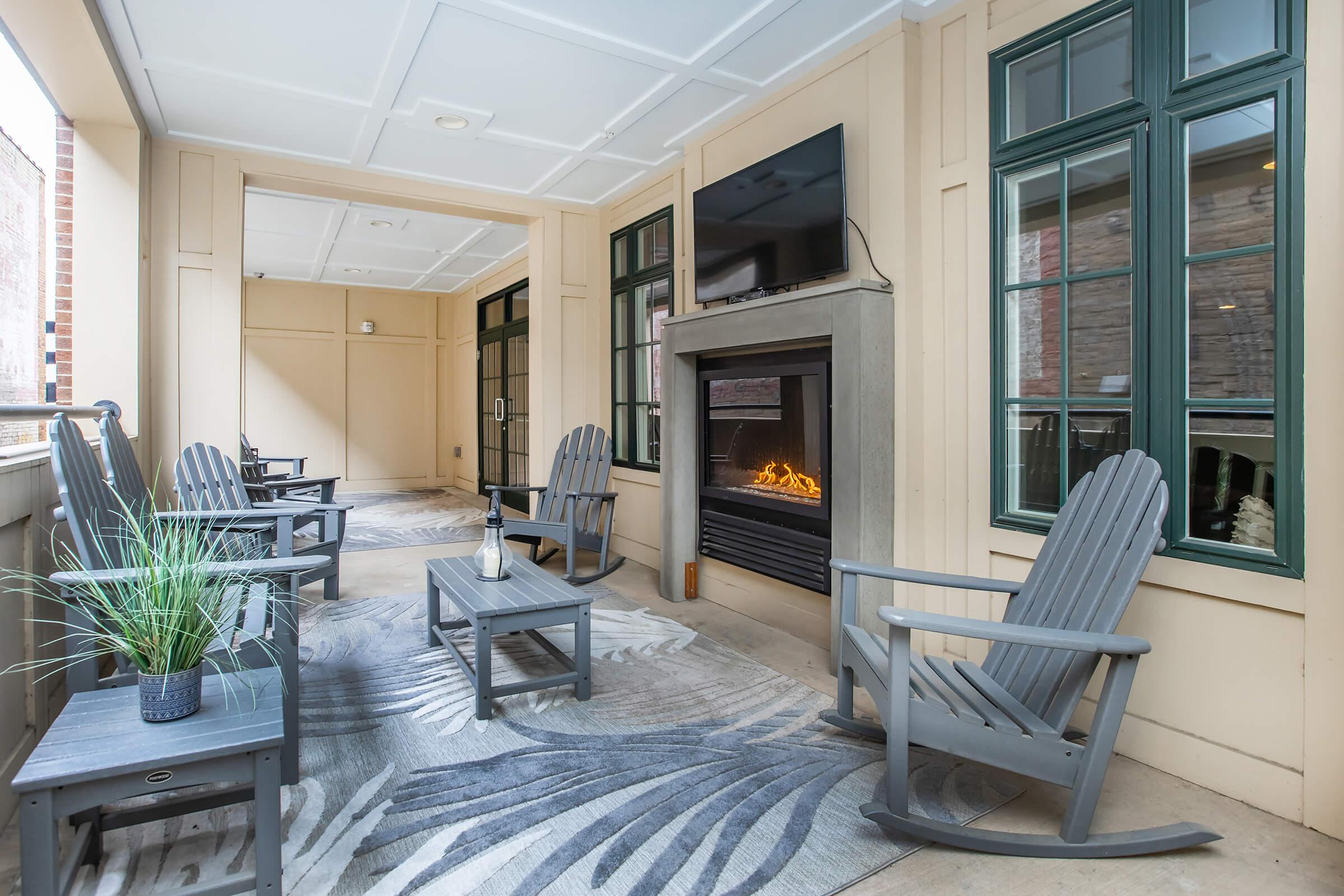 A cozy porch area featuring light-colored walls with large windows. There are four grey rocking chairs arranged around a coffee table, and a modern fireplace is mounted on the wall. A decorative rug adds a touch of warmth, and a small plant enhances the inviting atmosphere.