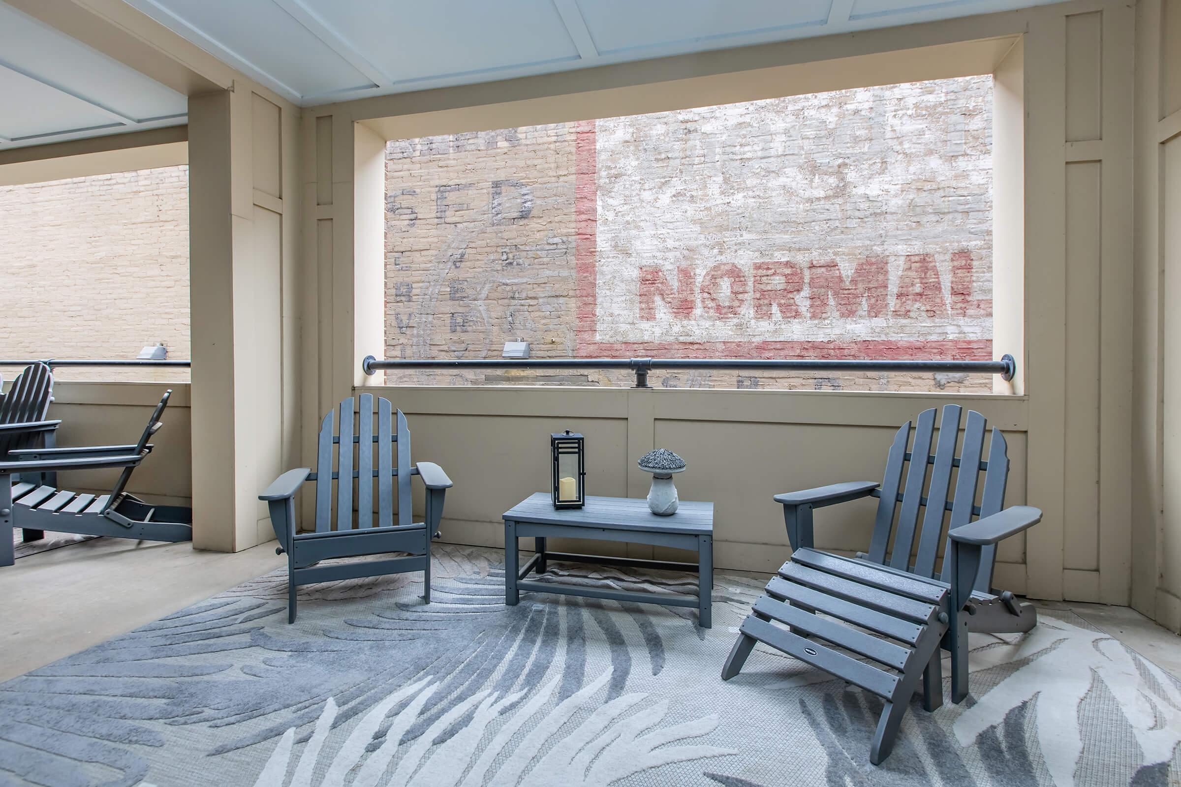 A cozy outdoor seating area featuring two blue Adirondack chairs and a small coffee table, set against a textured wall with faint vintage advertisements. A lantern and decorative mushroom figurine are on the table, and a palm-patterned rug adds a touch of style to the space. Natural light filters in from the window.