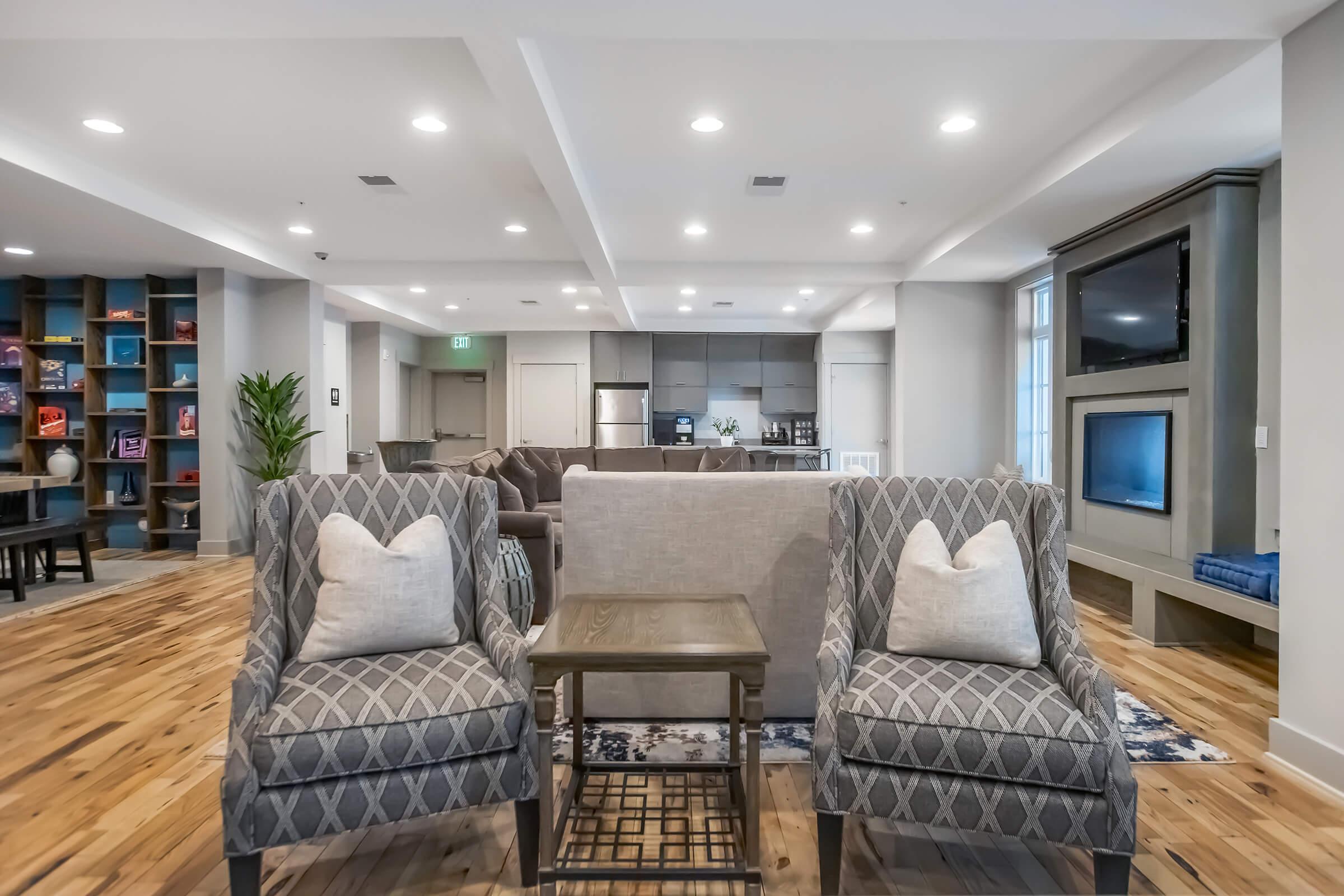 A cozy and modern lounge area featuring two patterned armchairs with pillows, a small wooden table between them, and a backdrop of a stylish living space with a sofa, TV, and kitchenette. Bright overhead lights illuminate the wooden flooring and contemporary decor.