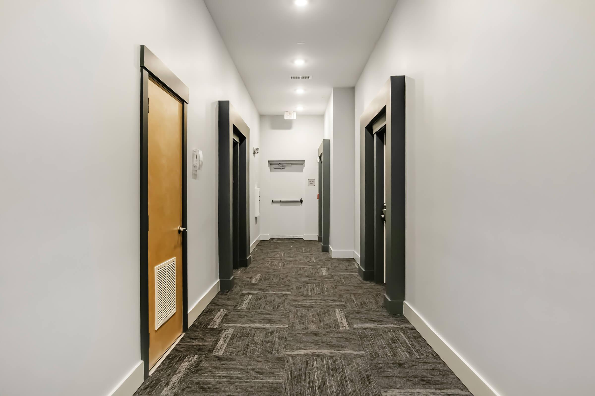 A well-lit, modern hallway featuring several doors on either side. The walls are painted white with dark-colored door frames. The flooring is covered in carpet with a textured pattern. There are ceiling lights providing illumination, and a fire exit sign is visible on the wall.