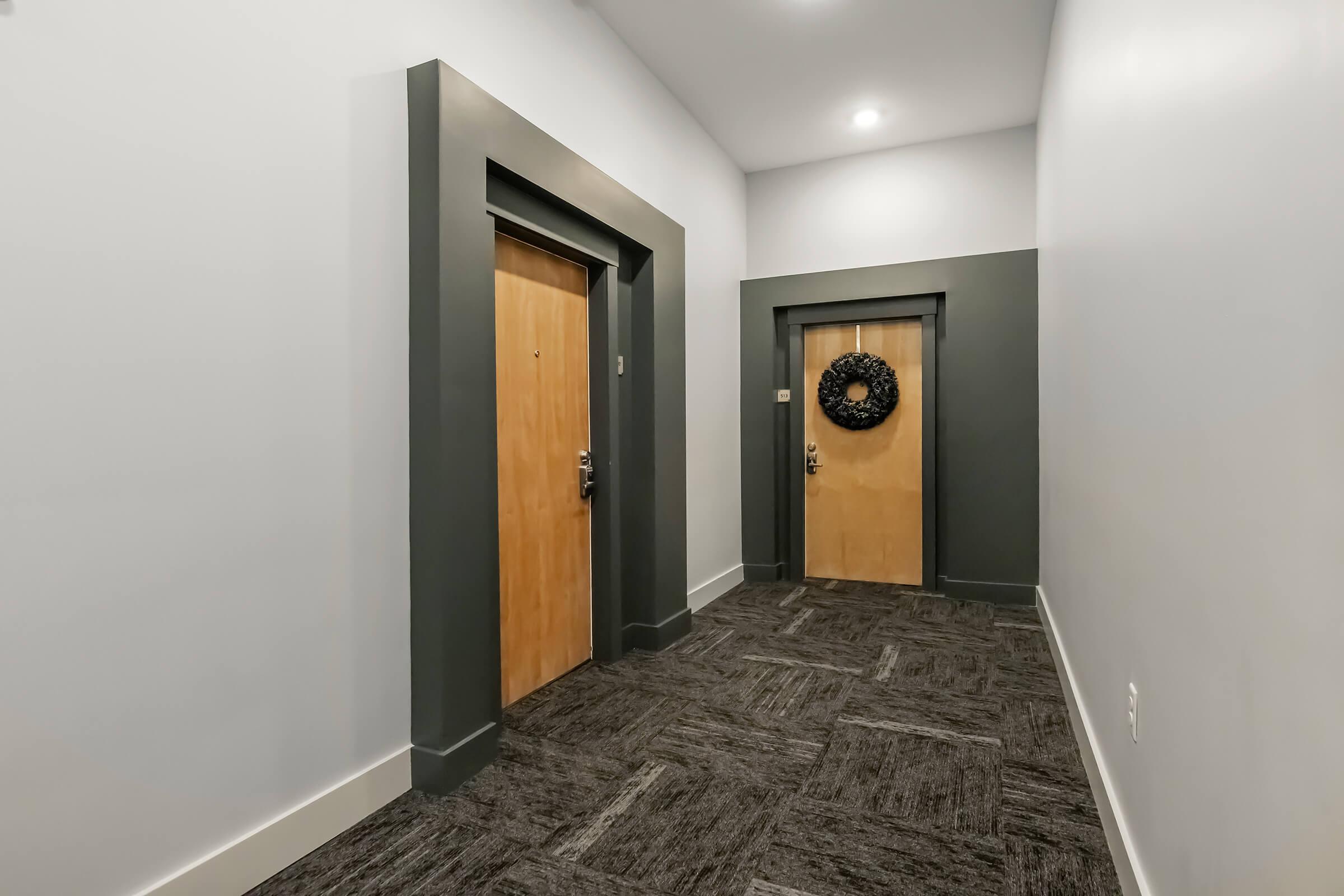 A modern hallway with two wooden doors, each with a simple gray frame. One door has a decorative black wreath, while the other is unadorned. The walls are painted light gray, and the flooring is dark textured carpet. Soft overhead lighting illuminates the space, creating a welcoming atmosphere.