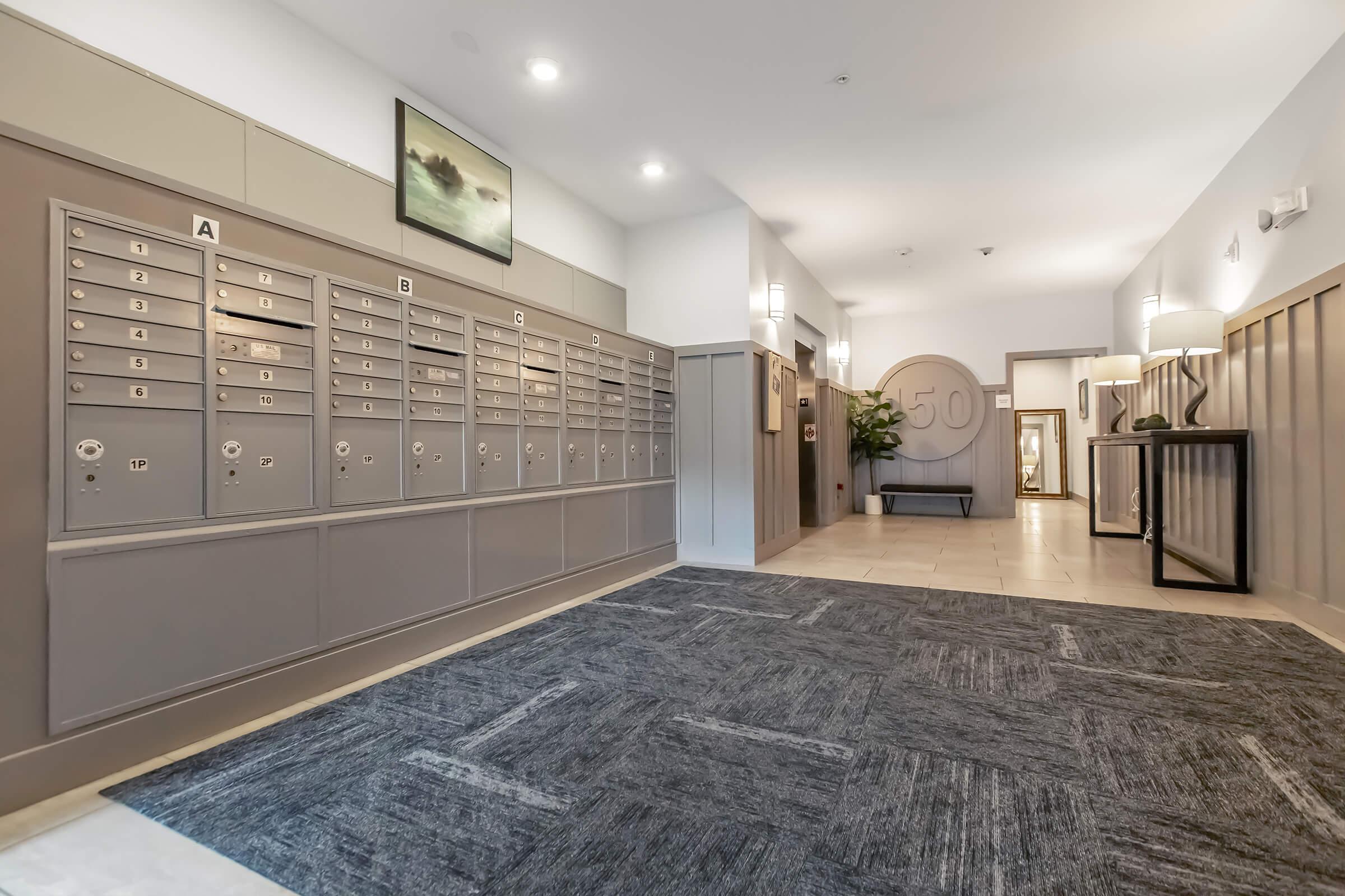 A modern apartment building interior featuring a mailroom. On the left, a row of secured mailboxes labeled A to D. The floor is covered with a gray carpet, and the walls are a soft neutral color. To the right, there is a decorative element with a round design featuring the number 50, along with plants and tasteful lighting.