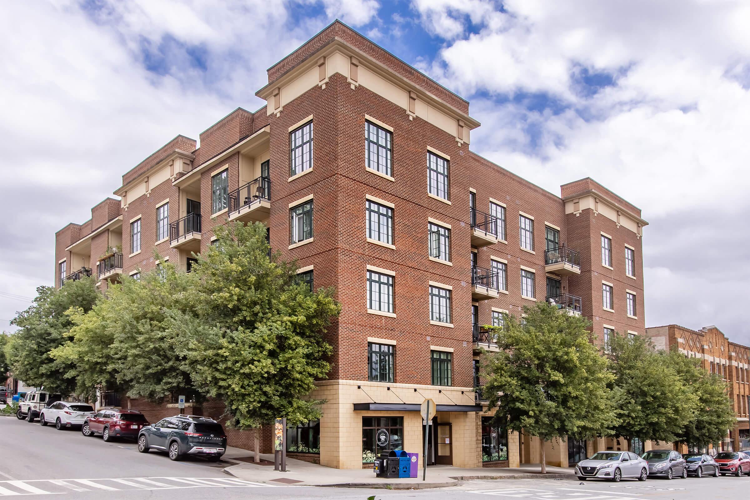 A modern brick apartment building with large windows and balconies, situated on a tree-lined street. Several cars are parked along the curb, and the sky is partly cloudy. The surrounding area features green trees and a mix of residential and commercial buildings.