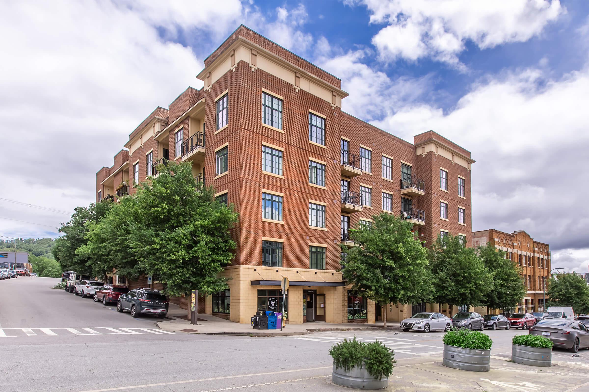 A brick apartment building with multiple stories and balconies, surrounded by trees. Cars are parked along the street in front, and there are large planters with greenery. The sky is partly cloudy, creating a vibrant atmosphere. The building is situated on a slight incline, adding depth to the scenery.
