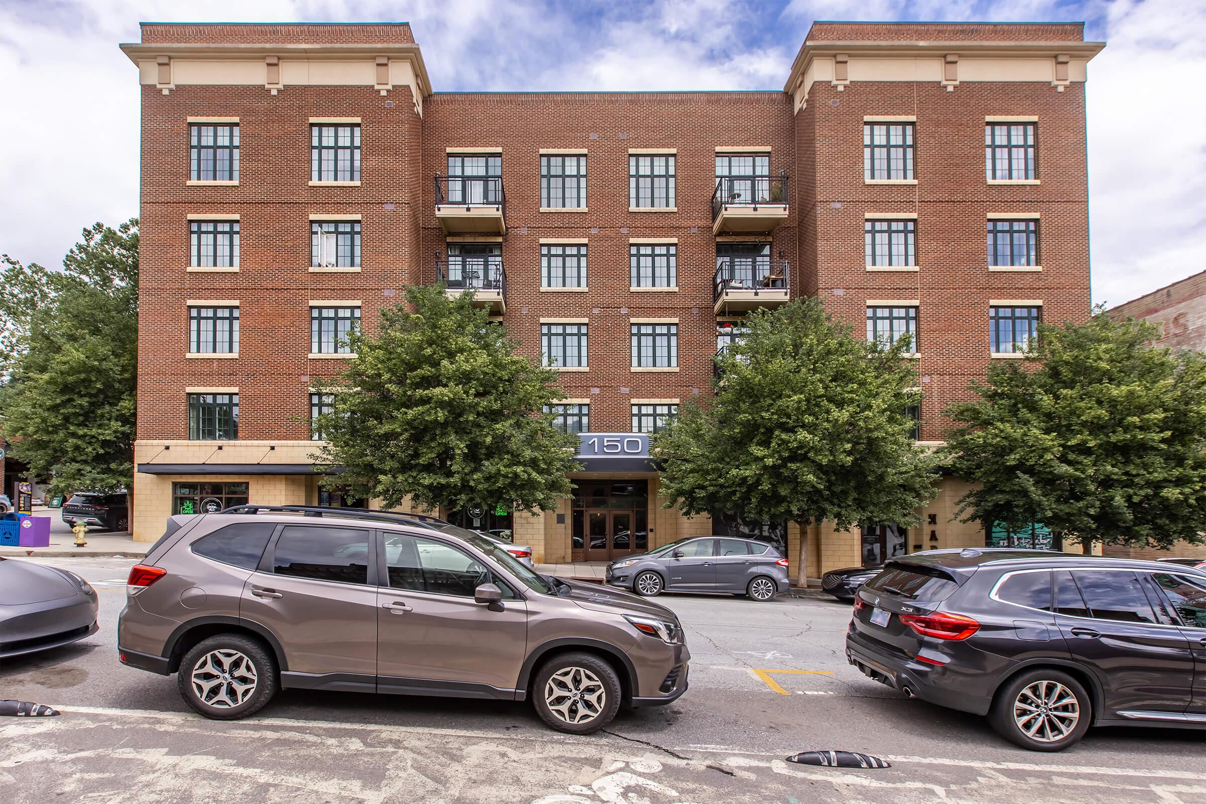A four-story brick building with large windows, balconies, and a central entrance labeled "150." Several trees line the sidewalk, and parked cars are visible in front of the building. The sky is partly cloudy, adding to the urban setting.