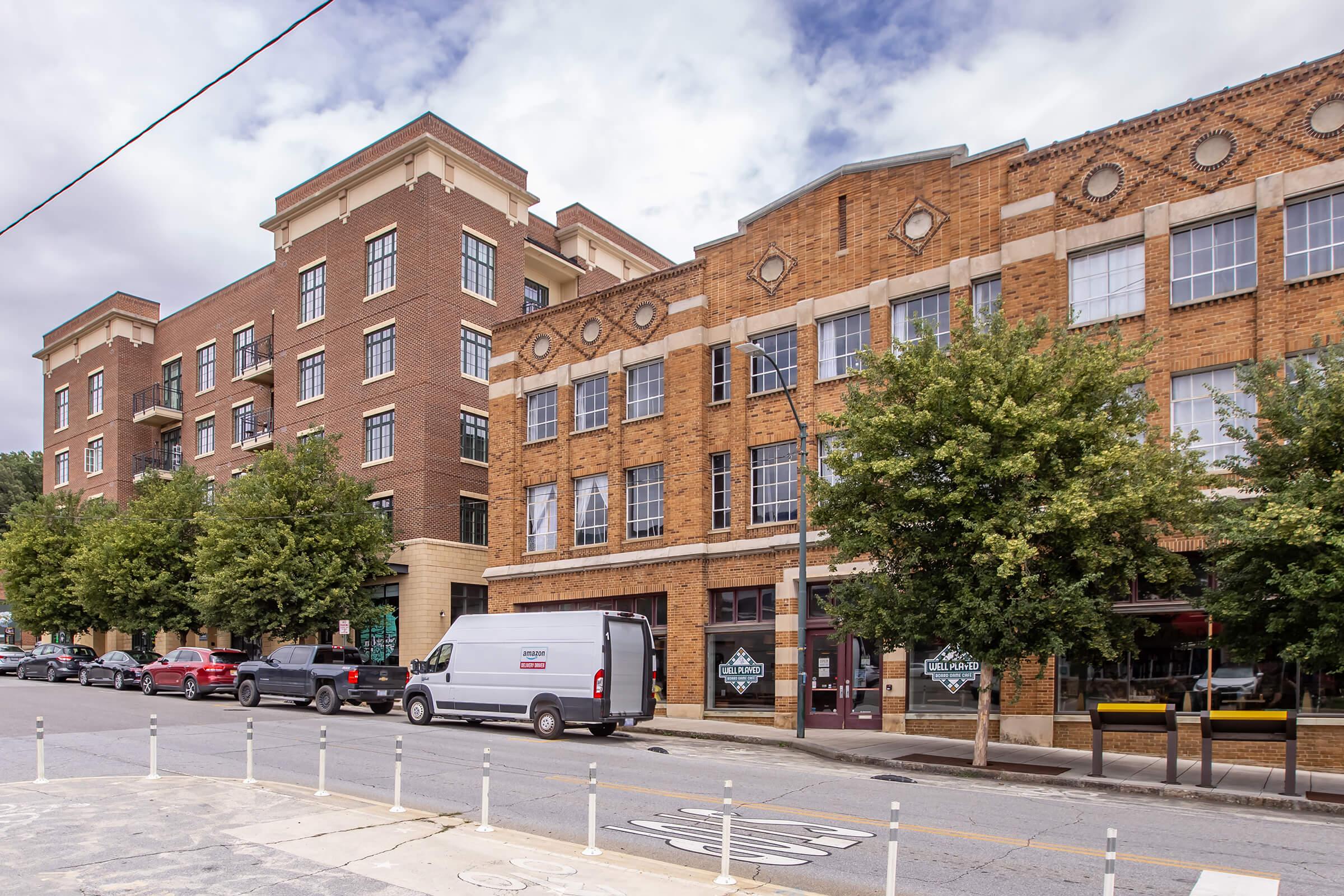 A street view of a city block featuring a mix of modern and historic brick buildings. The foreground shows parked cars along the road, and several trees lining the sidewalk. Cloudy skies are visible above, creating a muted atmosphere. The buildings have distinct architectural details.