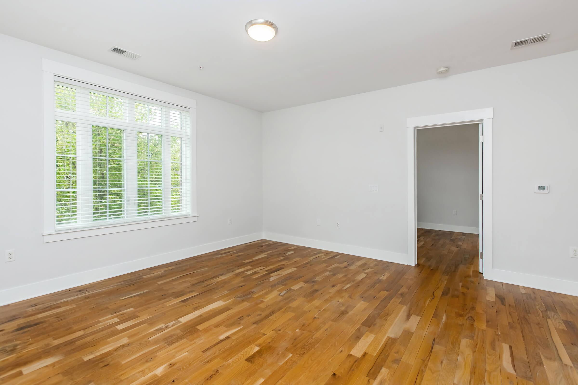 A spacious, empty room with hardwood flooring and white walls. There is a large window with blinds, providing natural light, and a doorway leading to another room. The ceiling features a central light fixture. The overall atmosphere is bright and minimalist.