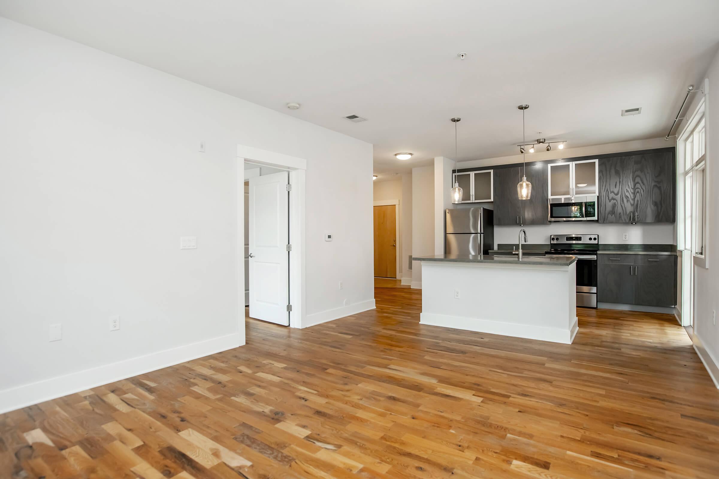 A spacious, modern apartment interior featuring an open-concept layout. The kitchen has sleek cabinetry, stainless steel appliances, and a breakfast bar. The living area has hardwood floors and natural light from large windows, with a white wall and a door leading to another room.