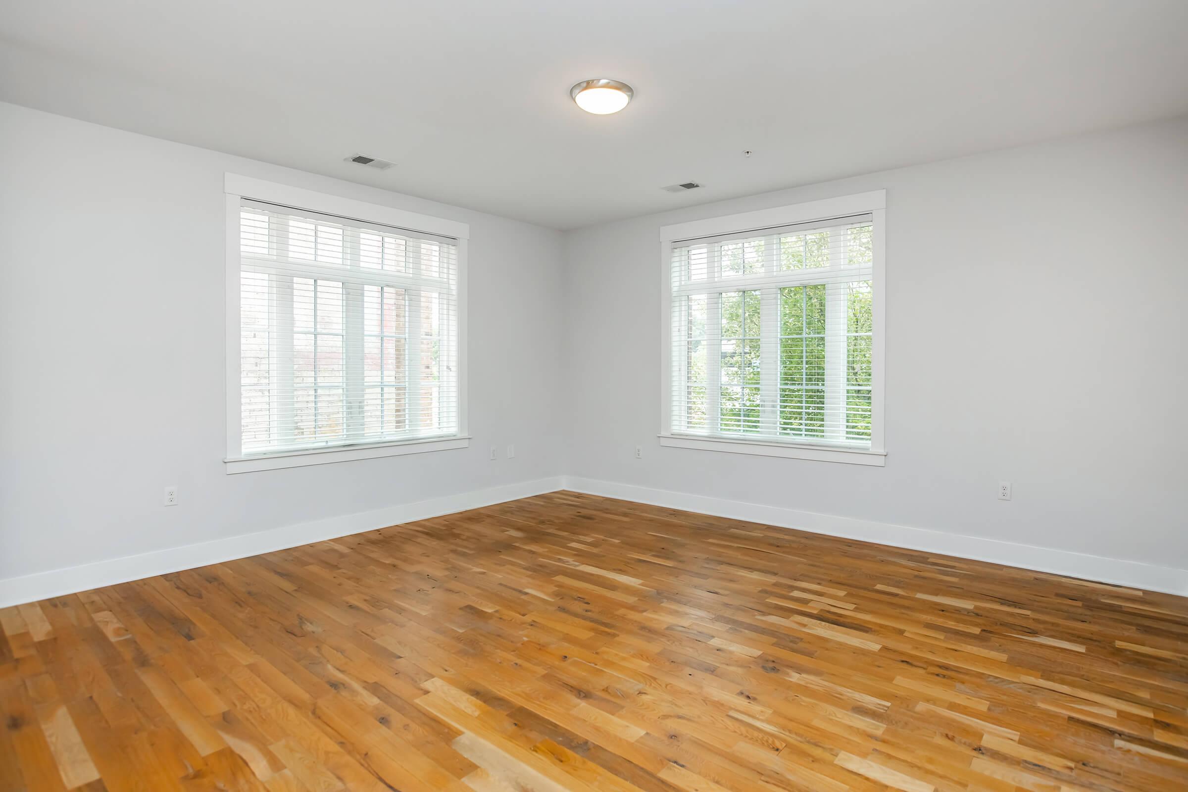 Empty room featuring light-colored walls, large windows with white shutters allowing natural light, and hardwood flooring. The space is minimalistic with no furniture, creating a bright and airy atmosphere.
