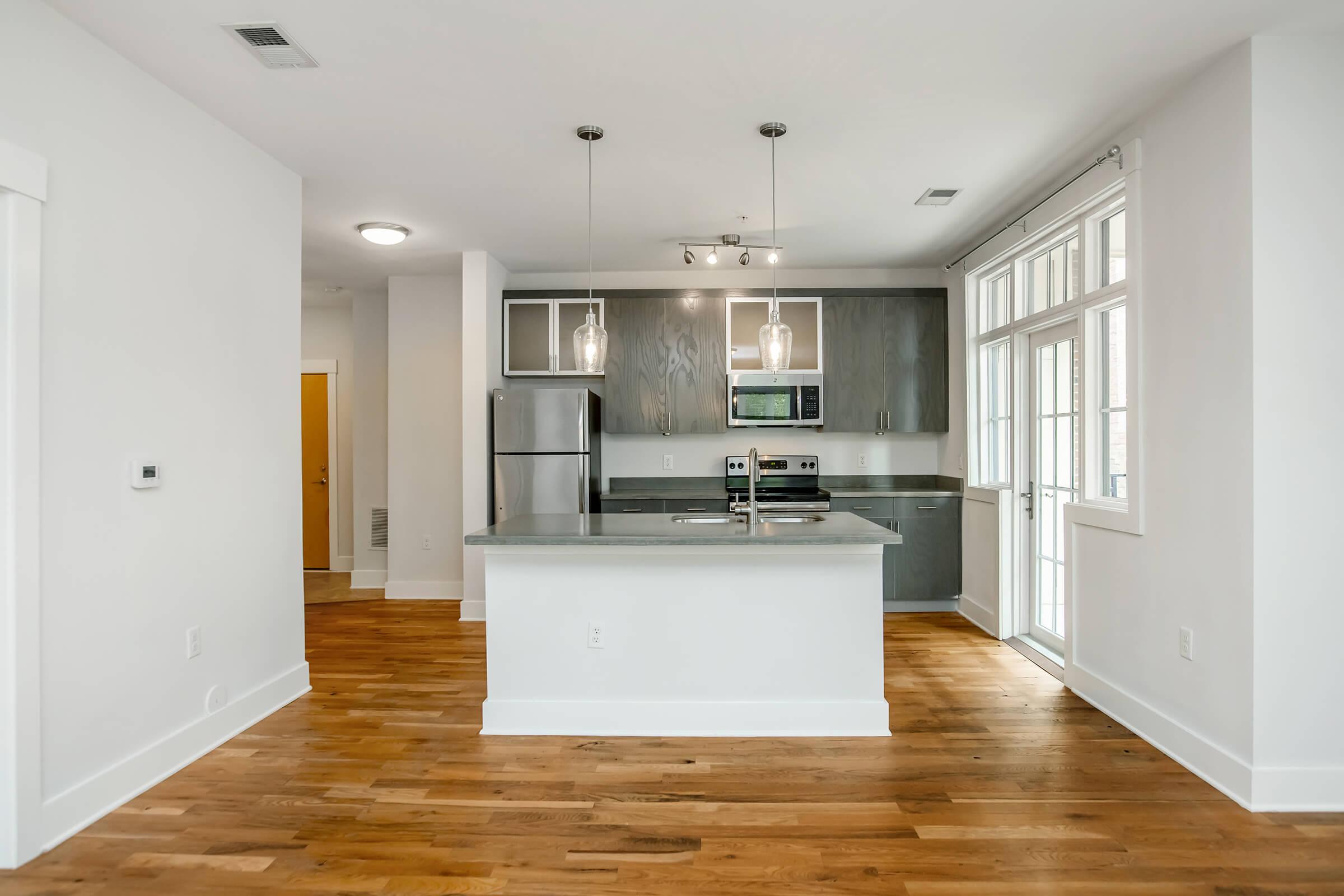 Modern kitchen featuring gray cabinetry, stainless steel appliances, and a large island with pendant lighting. The open layout includes hardwood flooring and windows allowing natural light. A door leads to an outdoor area. The design combines contemporary elements with functional space.