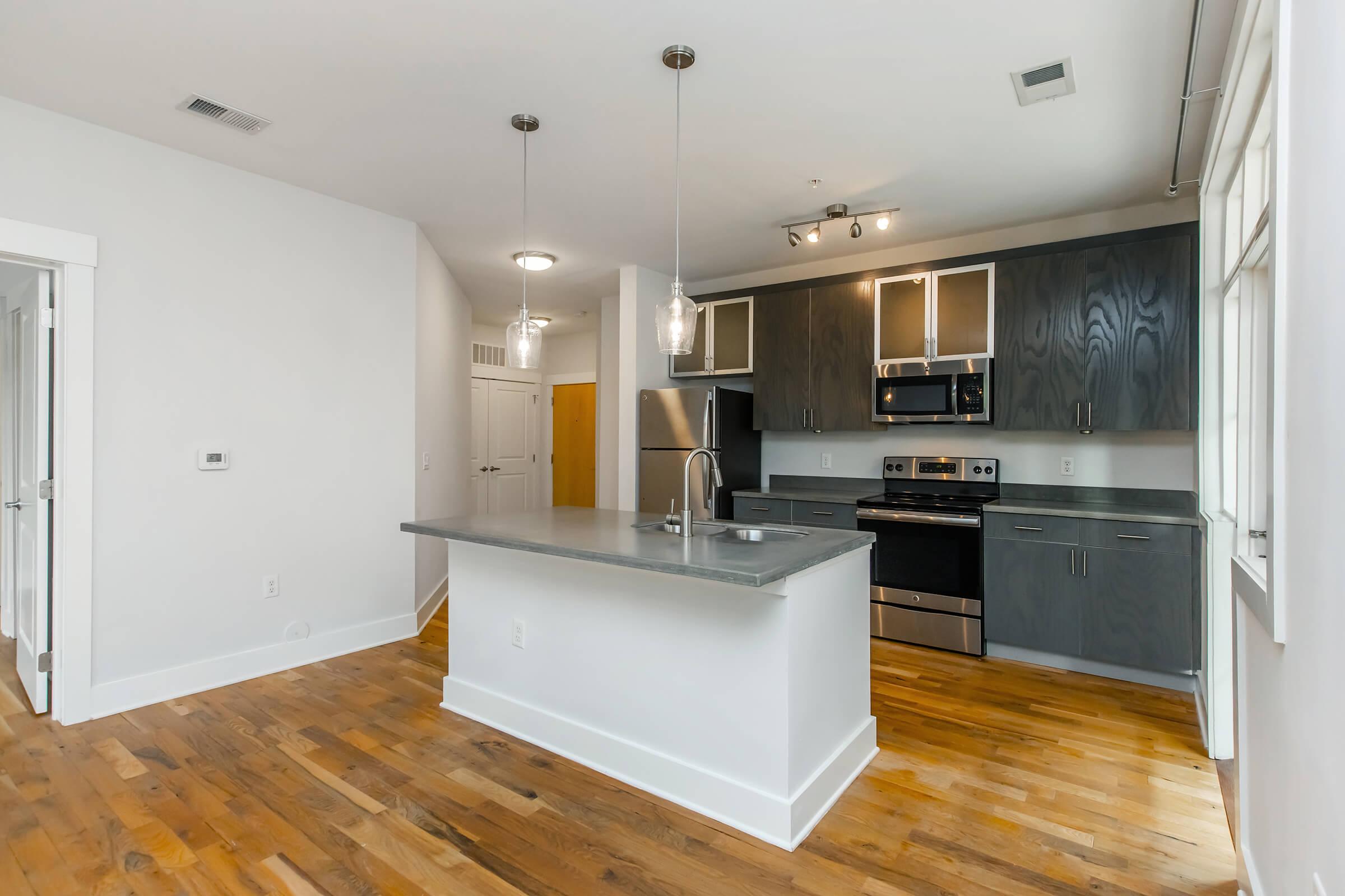 Modern kitchen featuring a central island with a stainless steel sink, dark cabinetry, and stainless steel appliances, including an oven, microwave, and refrigerator. The space has wood flooring and large windows, allowing for natural light. The design is sleek and contemporary, creating a welcoming atmosphere.