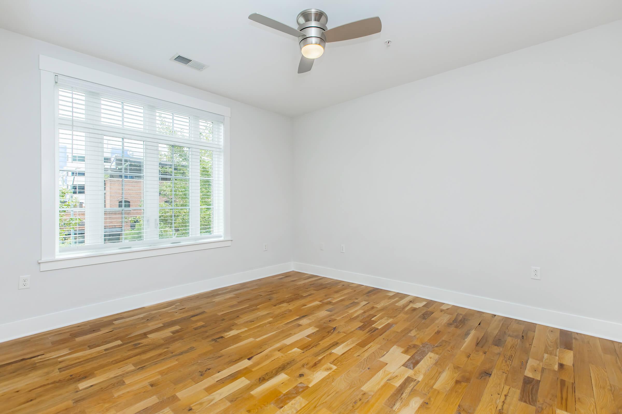 A spacious, empty room with light gray walls and hardwood flooring. A large window with white blinds allows natural light to flood the space, and a modern ceiling fan is visible. The room is designed for versatility and could serve as a bedroom, study, or living area.
