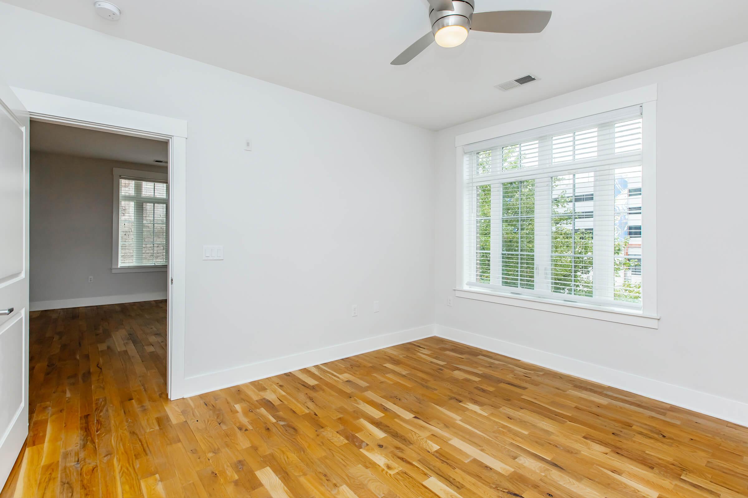 A bright, empty room with white walls and large windows adorned with blinds. The floor is made of polished wood, and a light ceiling fan is installed. A doorway leads to another room, suggesting an open layout in a modern living space. Natural light streams in from the windows, enhancing the airy feel.