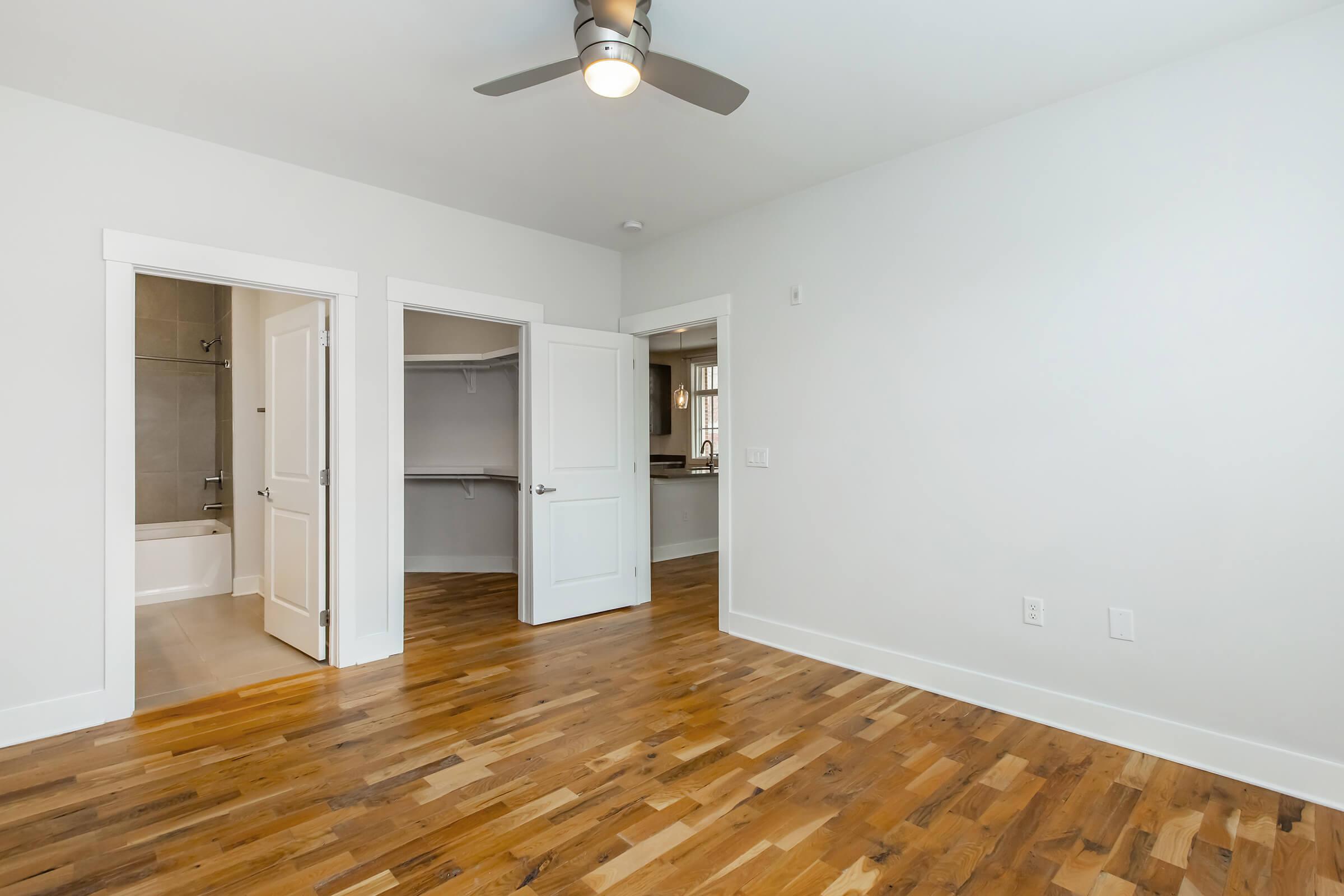 A bright, modern room with wooden flooring and light-colored walls. It features a ceiling fan, two open doors leading to a bathroom and a closet, and ample natural light, creating a spacious and inviting atmosphere.