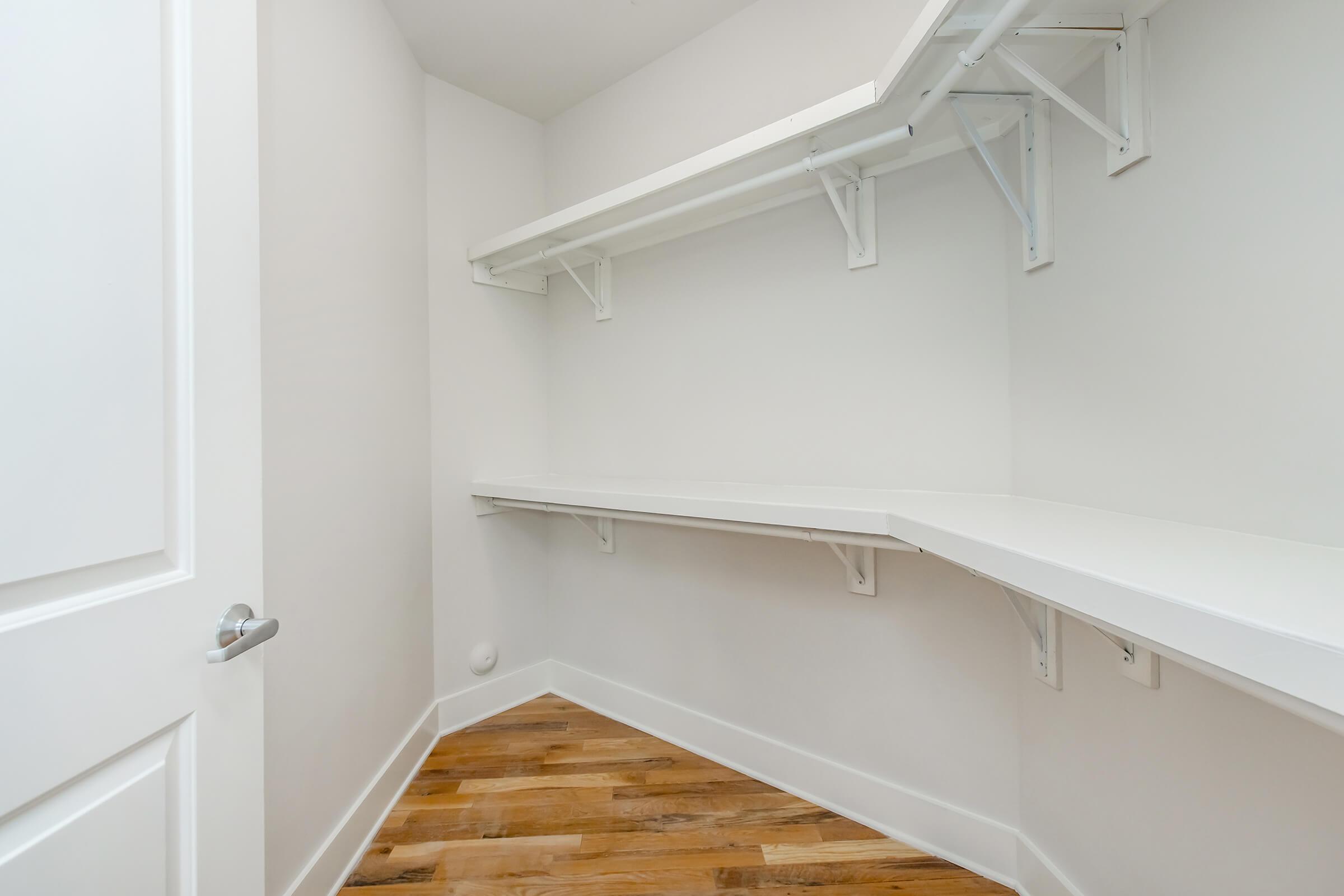 An empty walk-in closet featuring white walls and two floating shelves. The floor is made of hardwood, and there is a gray door partially visible on the left side. The overall space is bright and uncluttered.