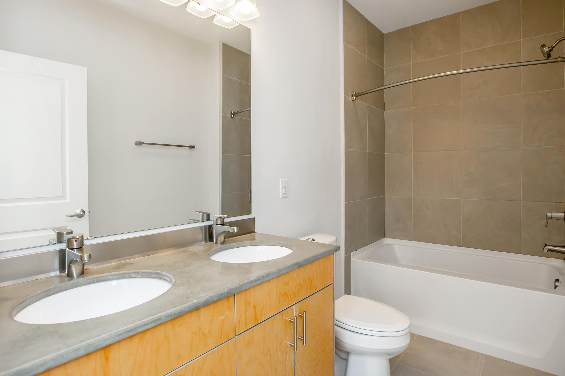 A modern bathroom featuring a double vanity with circular sinks and a large mirror above. The walls are tiled in neutral tones, and there is a bathtub with a shower head. A white toilet is visible, along with simple fixtures and a clean design. Natural light illuminates the space.
