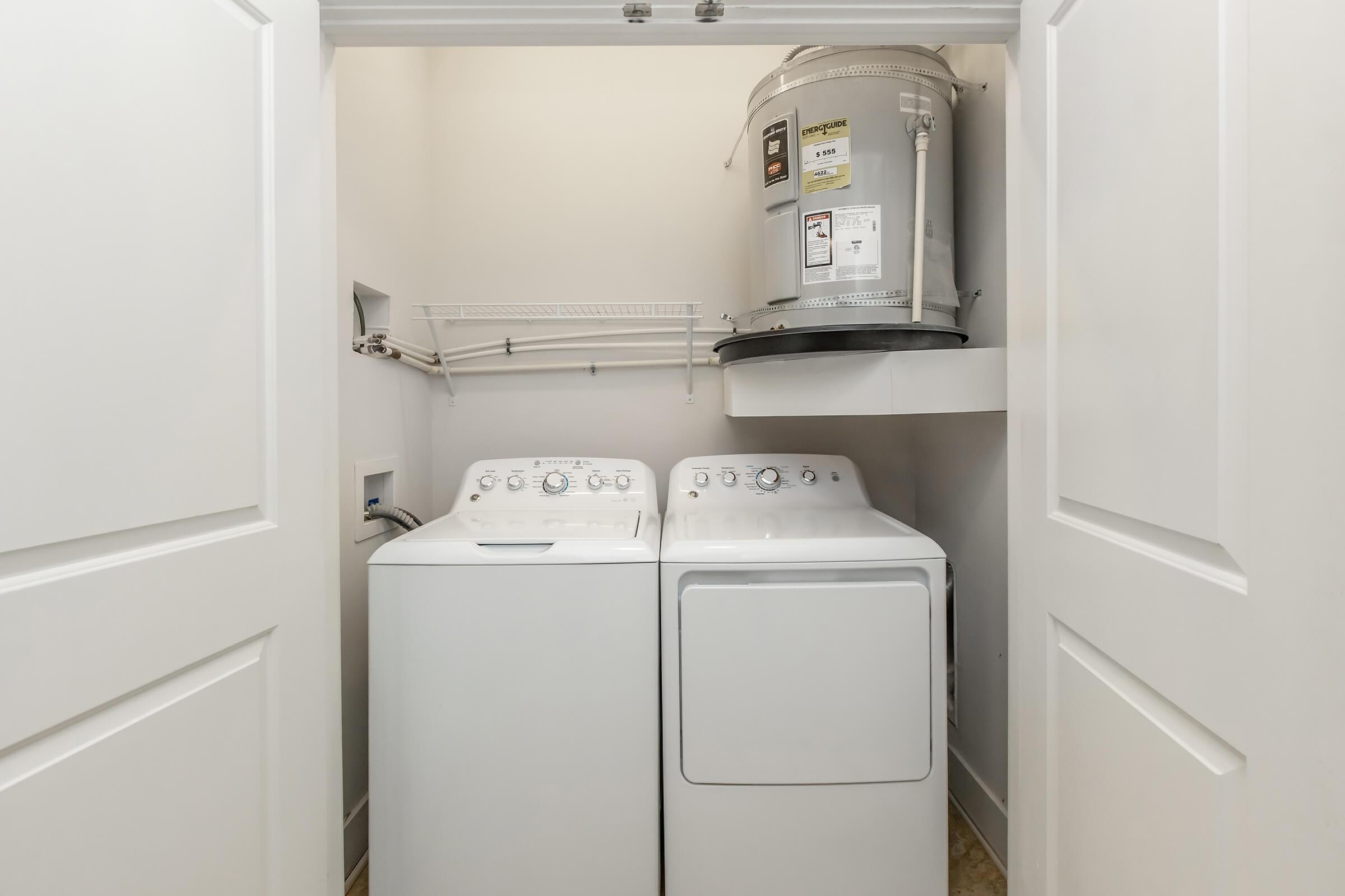A laundry room featuring a washing machine and a dryer side by side, with a water heater mounted on the wall above. The space is enclosed by white double doors, and there's a metal shelf above the appliances for additional storage. The floor is tiled and the walls are painted a light color.