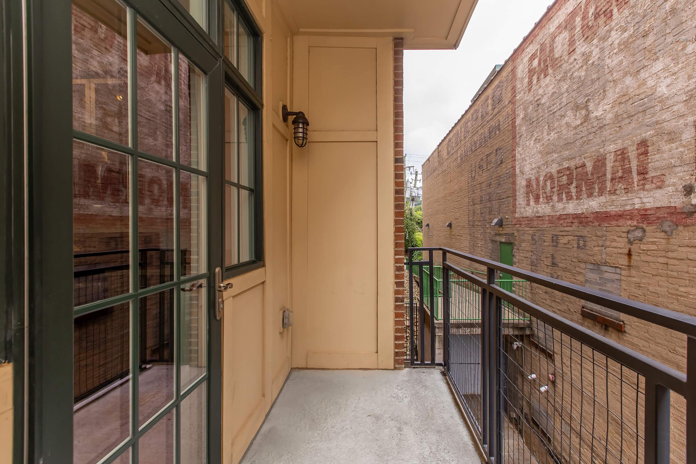 A narrow balcony with a metal railing, featuring a wall-mounted light fixture. To the left, there's a glass door leading inside, while the view shows a brick wall with faded advertisements. The balcony overlooks a narrow alleyway between two buildings, providing a small outdoor space in an urban setting.