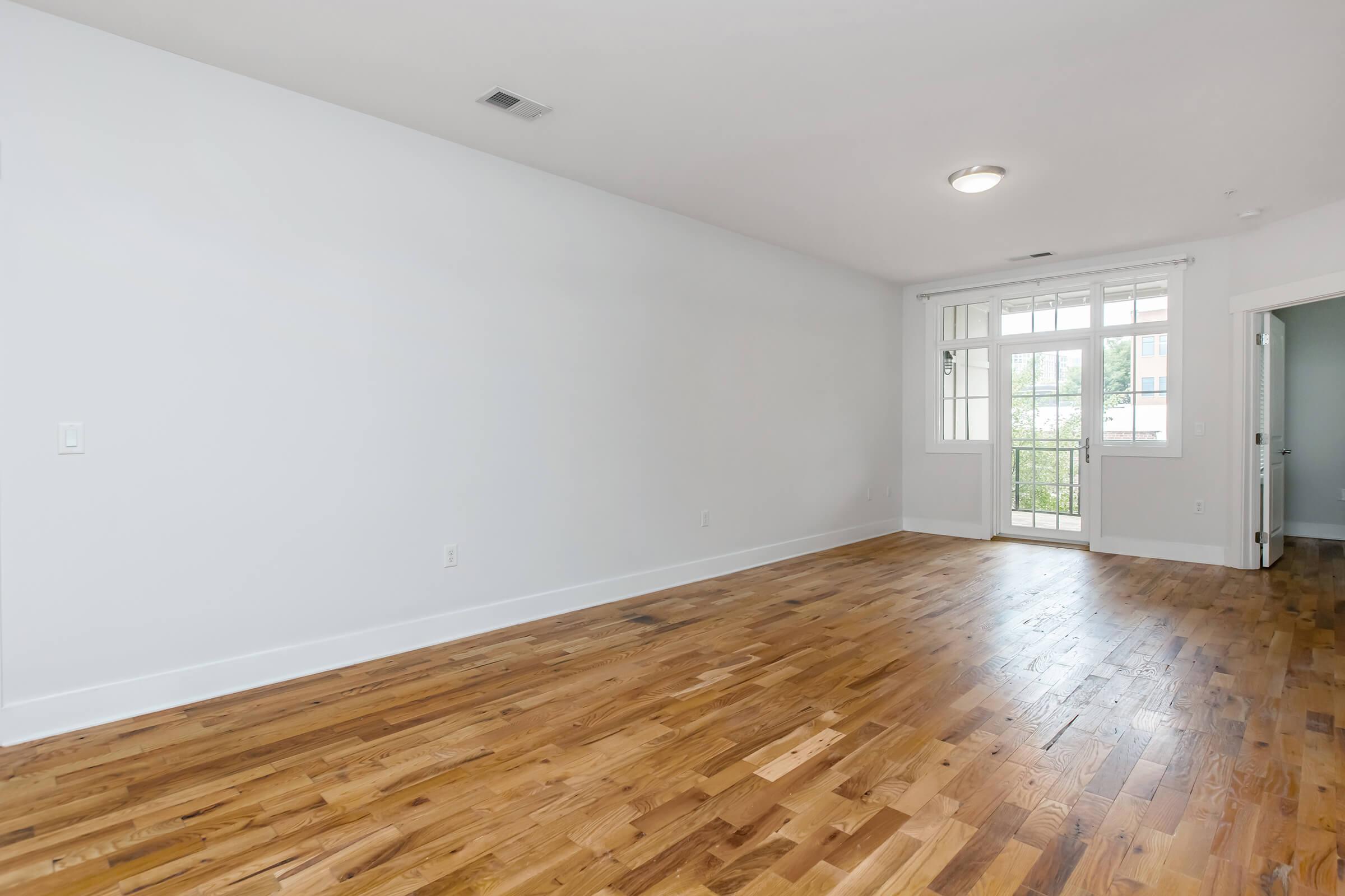 A spacious, empty room with hardwood flooring and white walls. A large window provides natural light, and there's a door leading to a balcony. The room is unfurnished and features a ceiling light, creating a clean and modern look.