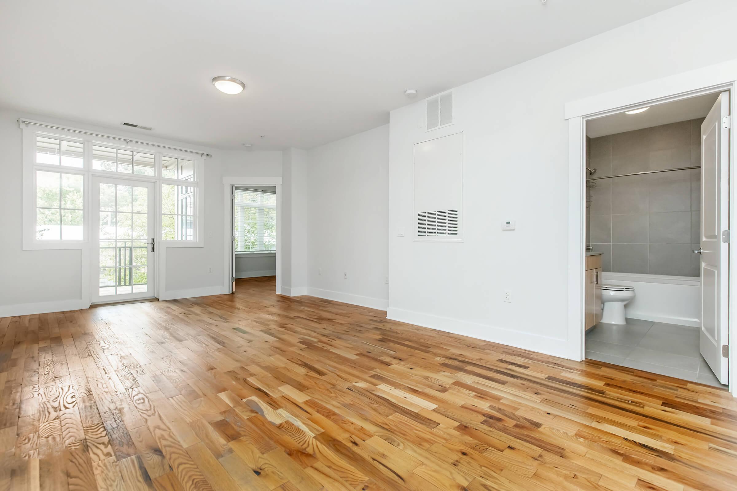 Spacious, well-lit room with dark wooden flooring, large windows, and a bathroom visible in the background. The walls are painted white, contributing to a bright and airy atmosphere. The layout features an open design, suitable for various furniture arrangements.