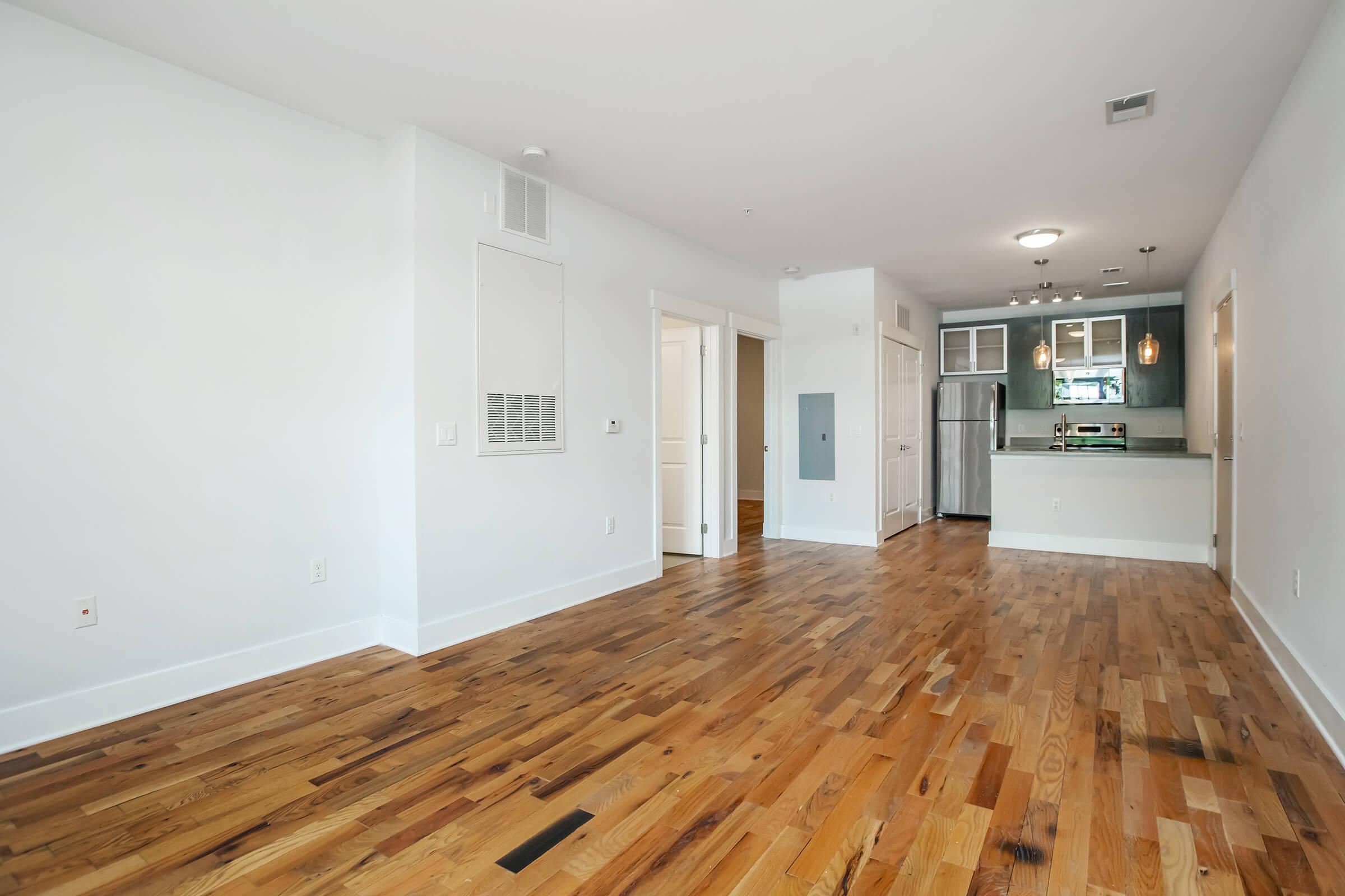 A spacious, modern apartment interior featuring hardwood flooring, white walls, and a kitchen area in the background. The kitchen has stainless steel appliances, cabinetry, and pendant lighting. The living area is well-lit with natural light and has a clean, minimalist aesthetic.
