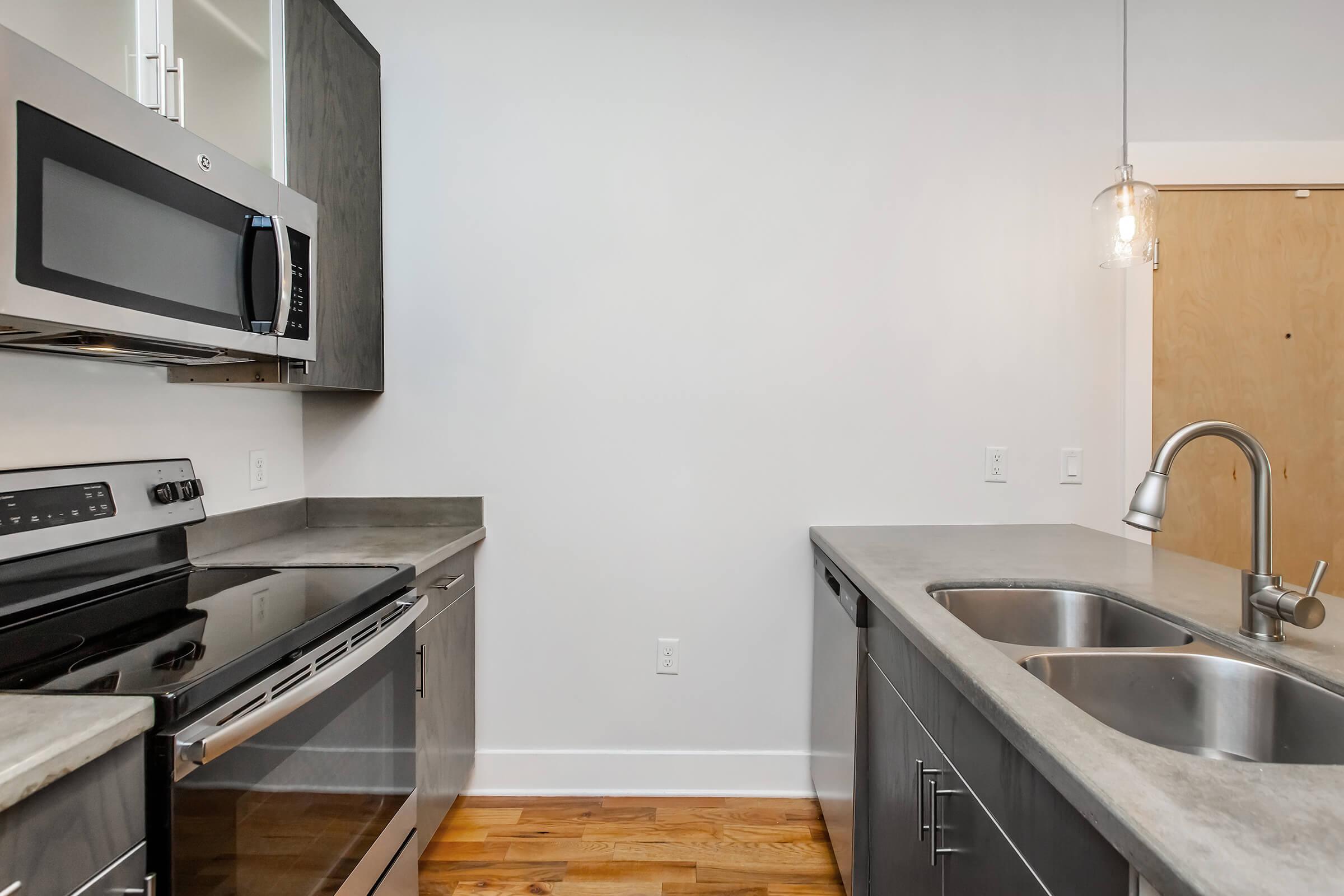 Modern kitchen featuring stainless steel appliances including an oven and microwave, concrete countertops, a double sink, and wooden flooring. There is a pendant light hanging above the sink area, and minimalist cabinetry in a sleek design. The walls are painted white, enhancing the contemporary aesthetic.