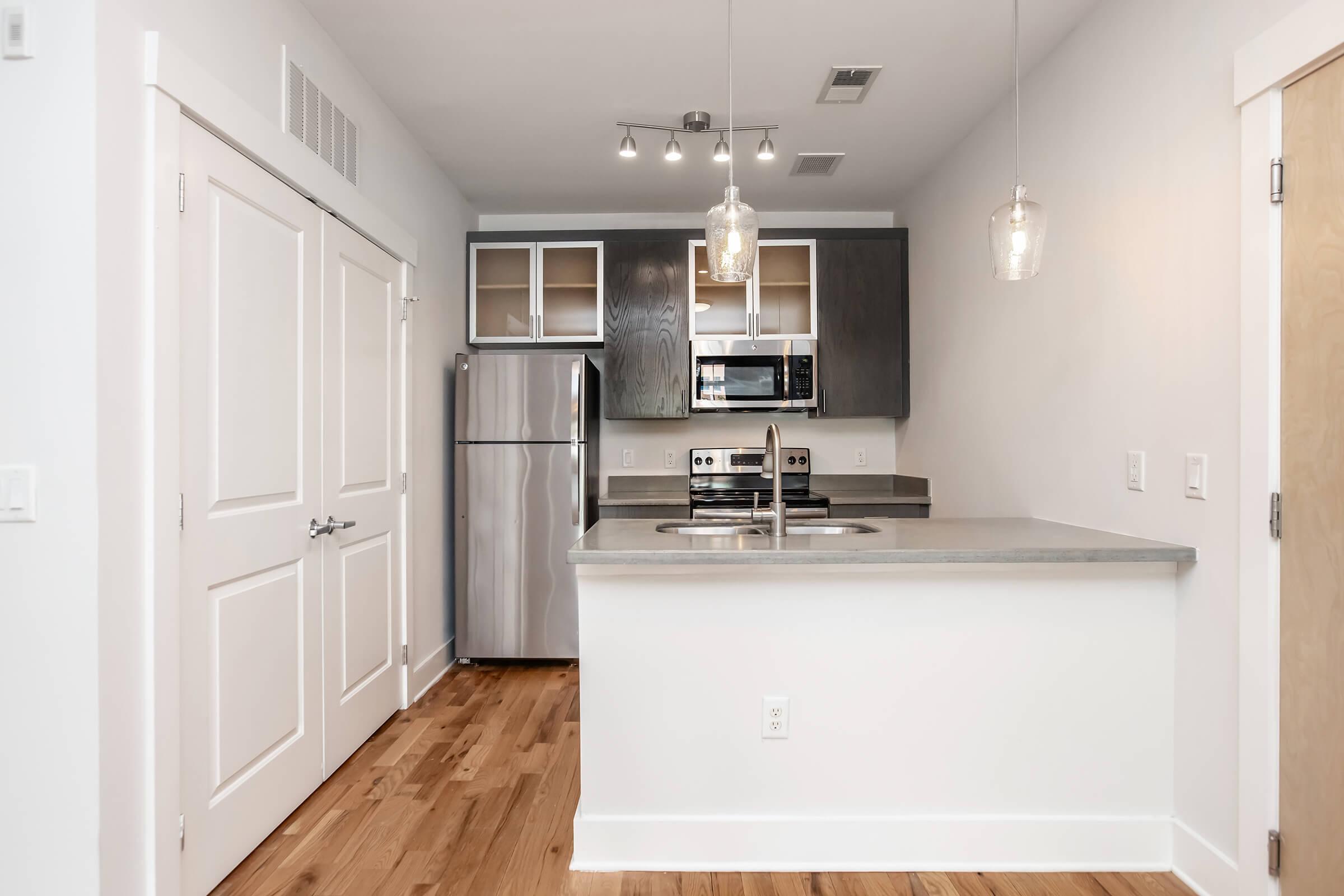 A modern kitchen featuring stainless steel appliances, including a refrigerator and oven, with gray countertops and a white backsplash. There are open shelves above the countertop, pendant lighting, and light wood flooring, creating a bright and contemporary atmosphere.