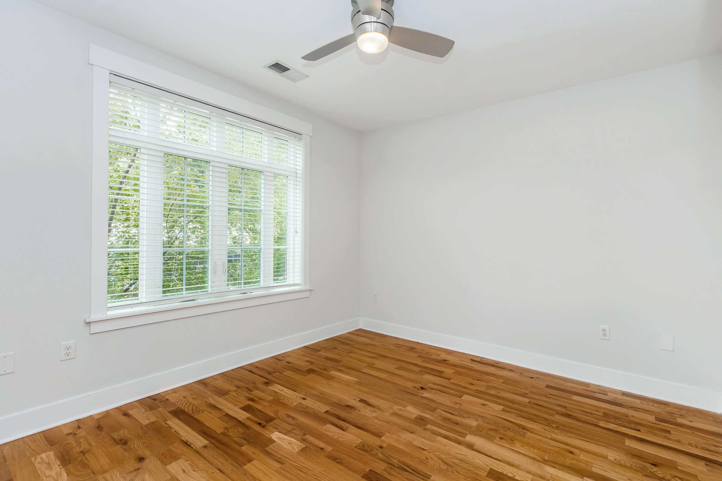 A bright, empty room with hardwood flooring and a ceiling fan. The large window with white blinds allows natural light to enter, illuminating the white walls. The space has a clean and minimalist design, perfect for various uses like a bedroom or office.