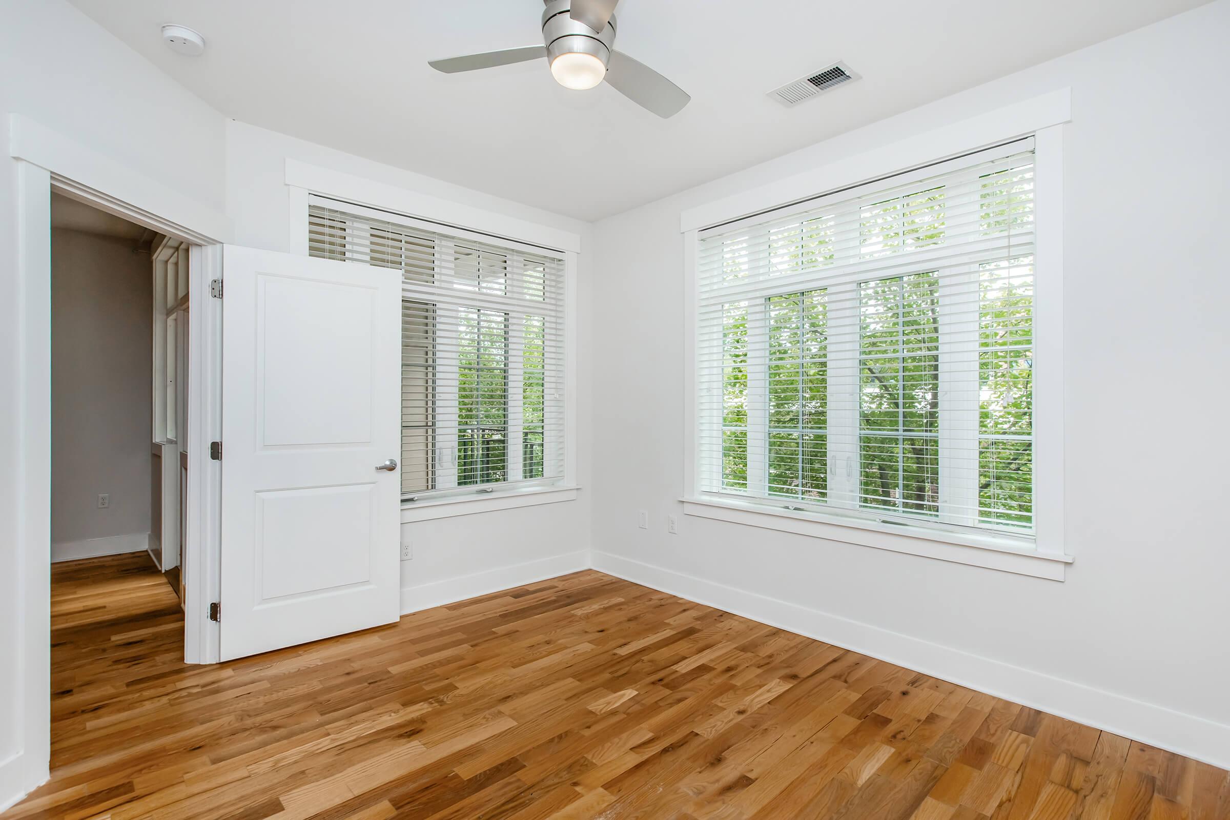 A bright, empty room featuring large windows with white blinds, hardwood flooring, and a ceiling fan. The walls are painted white, and a partially open door leads to another space, enhancing the airy feel of the interior.