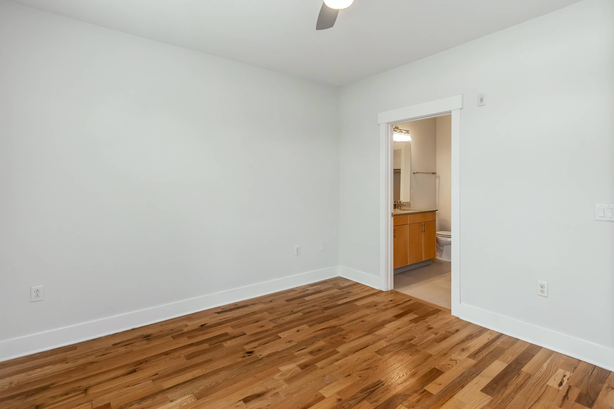 A bright, empty room with white walls and hardwood flooring. There's an open doorway leading to a bathroom area, visible through a second doorway. A ceiling fan is installed, adding to the modern aesthetic. The space is uncluttered, showcasing its potential for personalization.