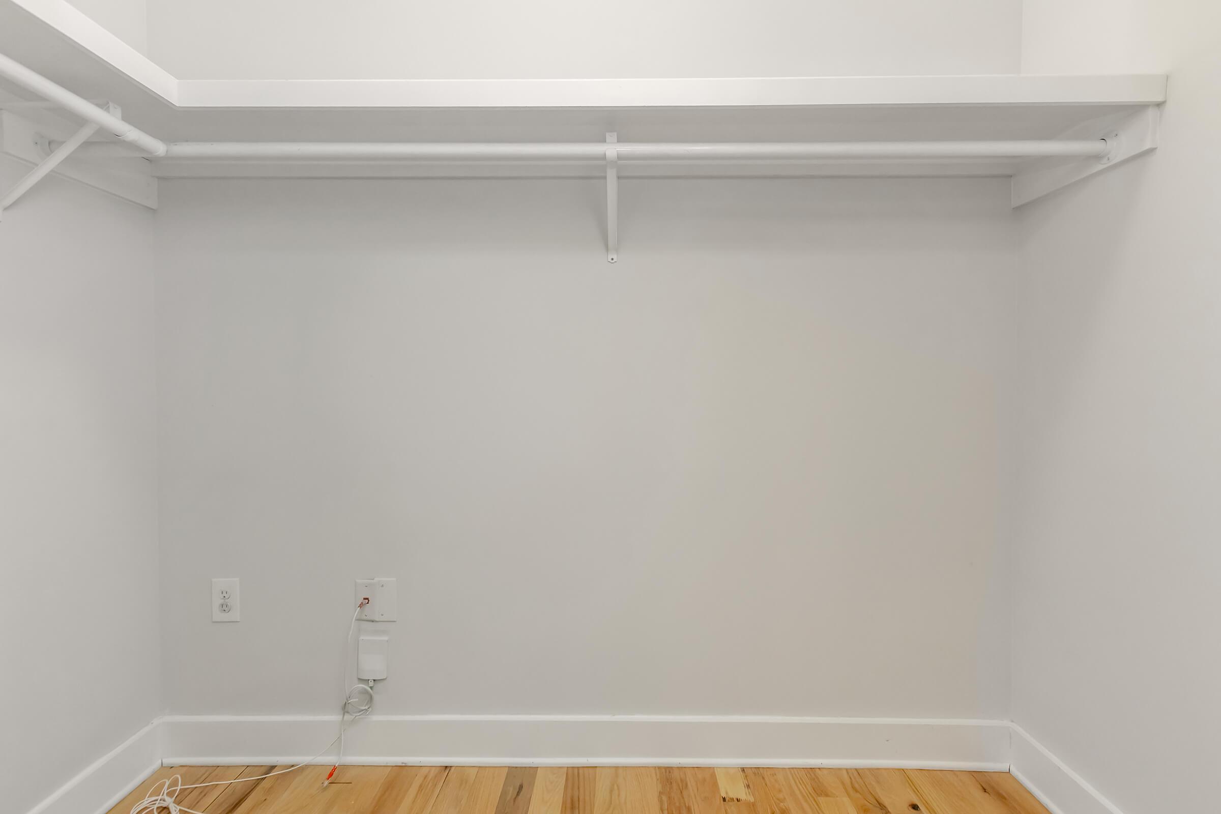Empty closet space with a white wall, a wooden floor, and a single shelf. There is a white hanging rod installed on the wall and a small electrical outlet with a visible cord. The overall appearance is clean and minimalistic, with no items stored in the closet.