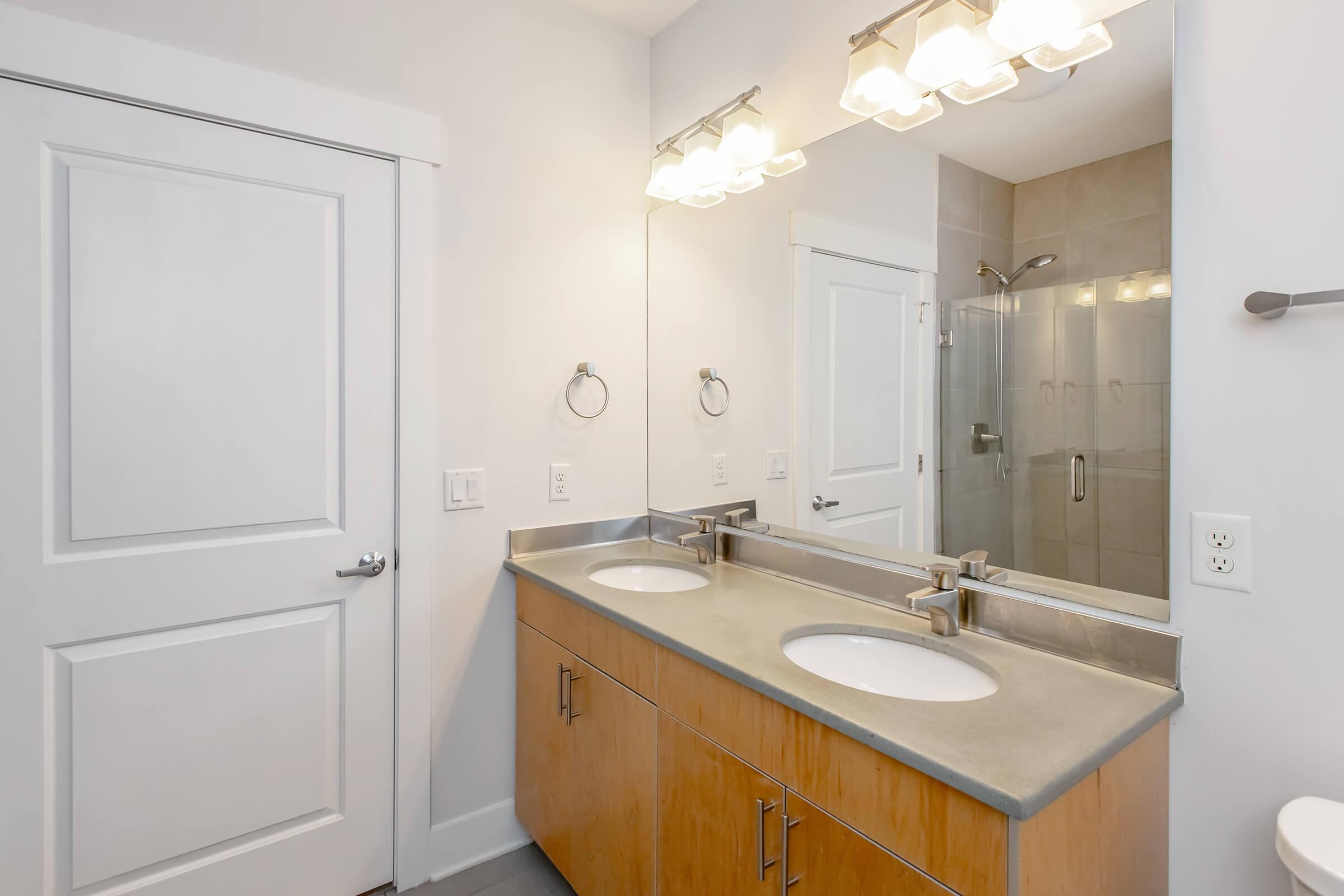 Modern bathroom featuring a double sink vanity with a concrete countertop, two mirrors, and a glass-enclosed shower. The decor includes light-colored walls and wood cabinetry, with a bright lighting fixture above the sinks. The space is well-lit and maintains a clean, minimalist aesthetic.