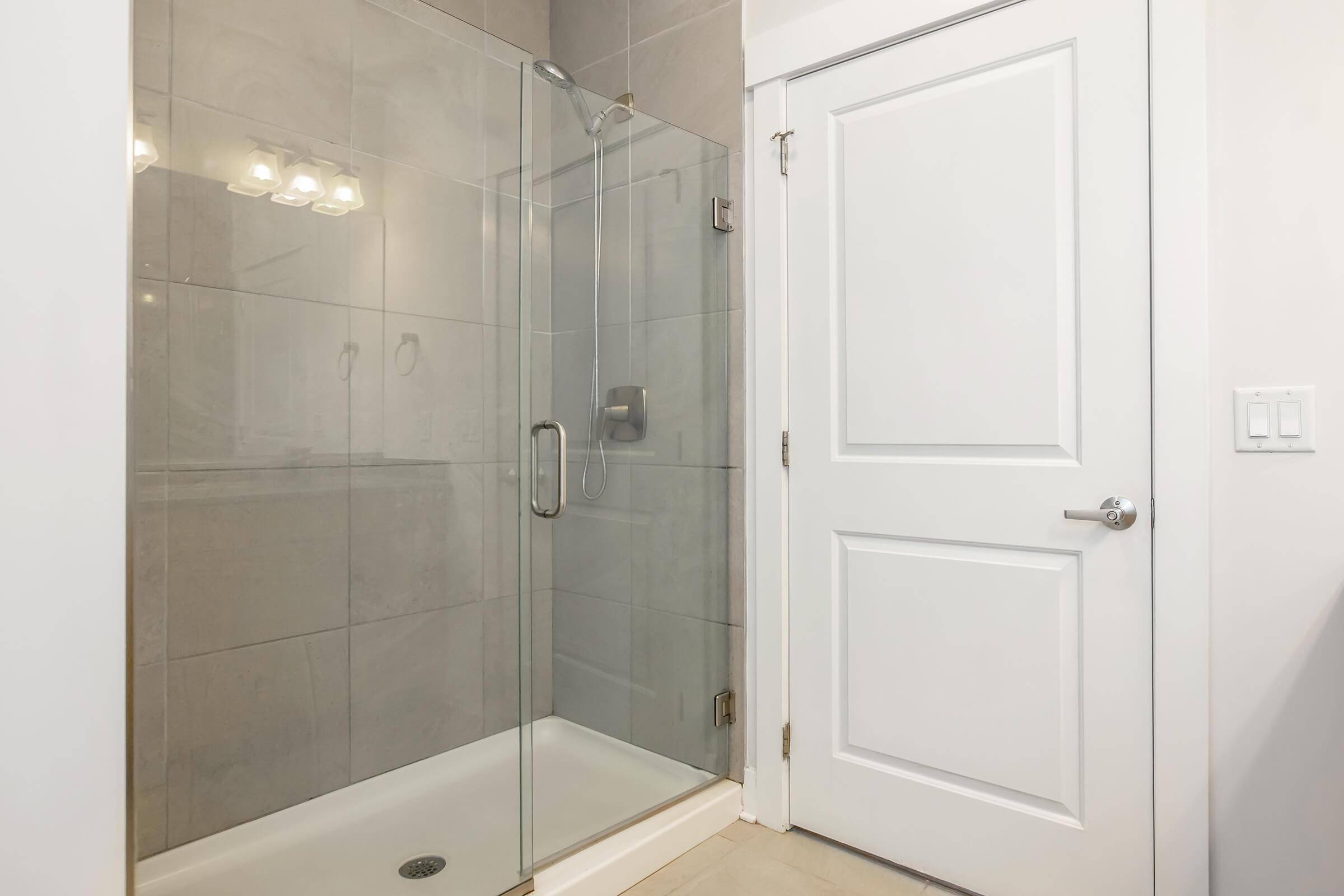 A modern bathroom featuring a glass shower enclosure with a chrome showerhead, gray tiled walls, and a white door. Soft lighting illuminates the space, highlighting the clean and contemporary design. The shower area is spacious with a tiled floor, creating an inviting environment.