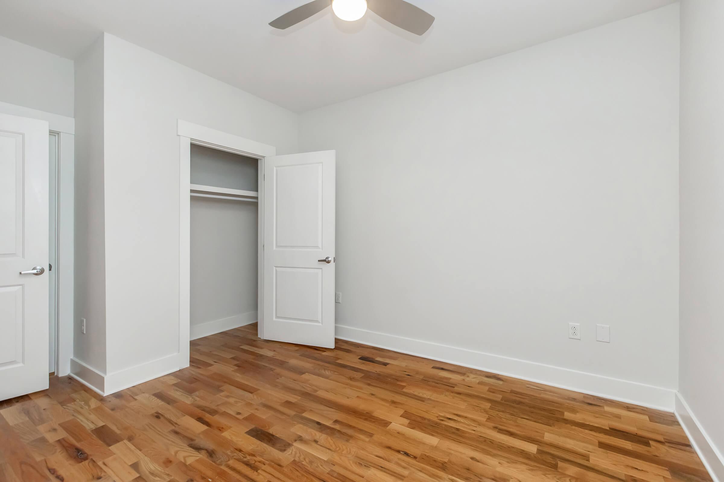 Empty room with light-colored walls and a smooth, wooden floor. A ceiling fan is installed, and there is an open closet with a white door on the left. Natural light illuminates the space, creating a clean and modern feel.