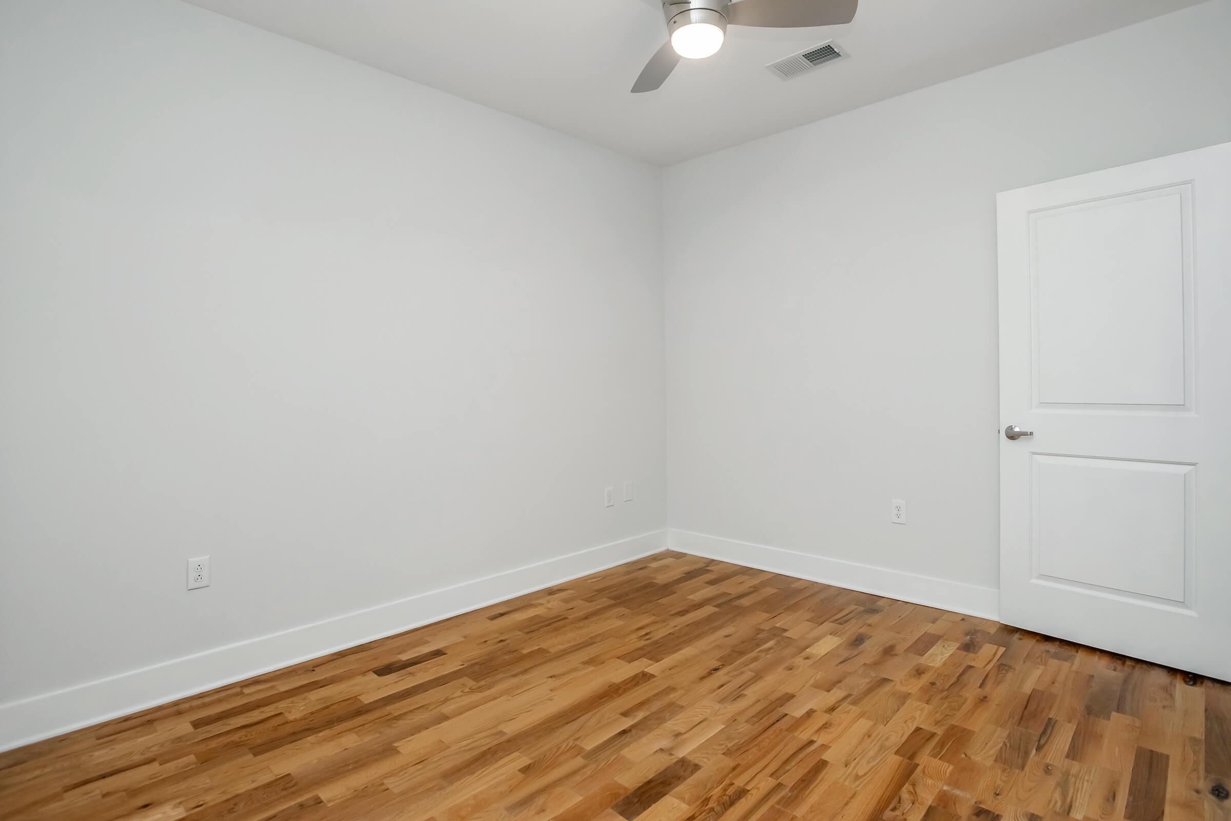 A spacious, empty room with light-colored walls and a hardwood floor. A ceiling fan is visible, and there is a closed white door on the right side of the image. The room is well-lit, suggesting a bright and airy atmosphere, suitable for various purposes like a bedroom or office.