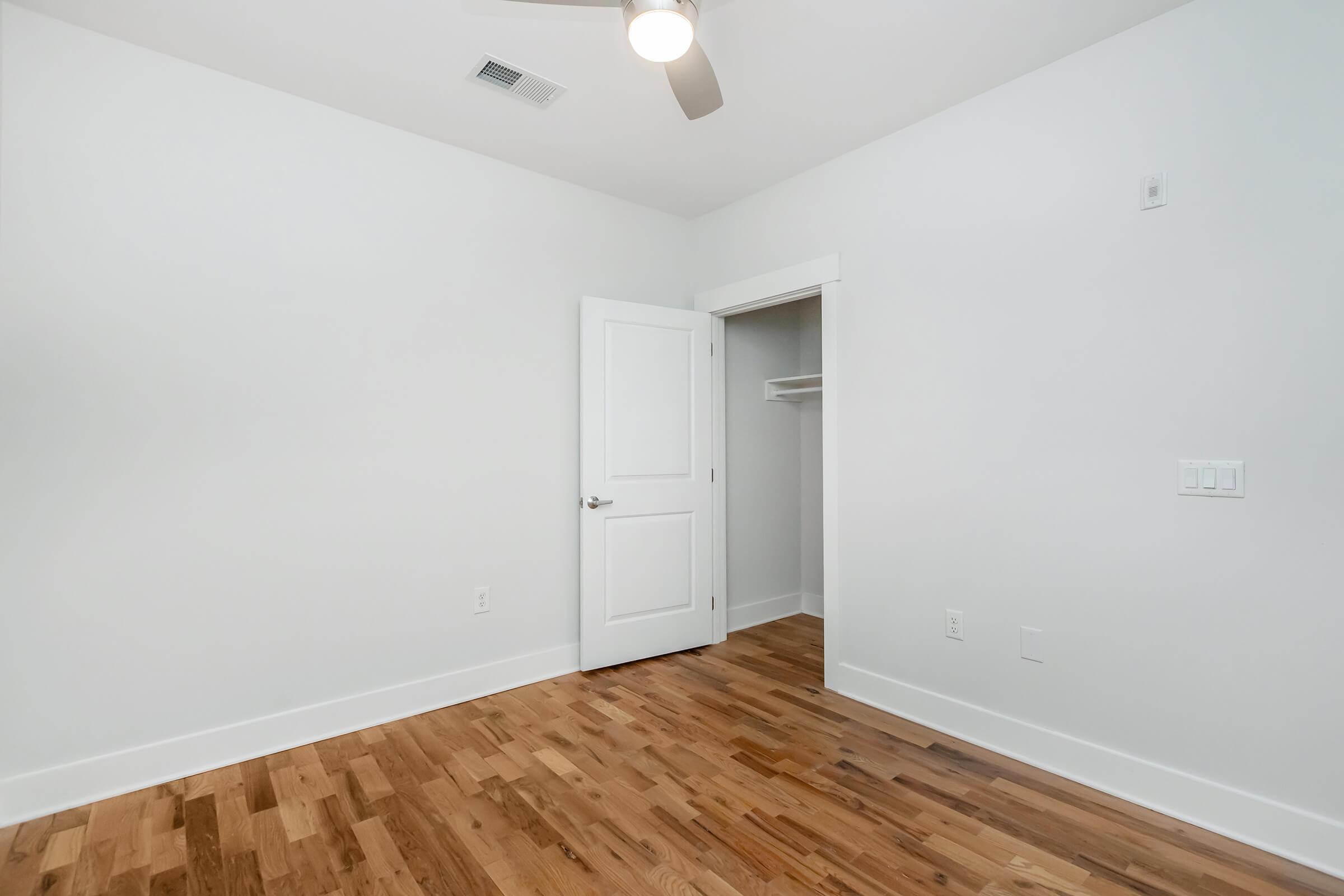 A bright, minimalistic room with white walls and a light wood floor. A door with a silver handle is slightly ajar, revealing a closet space inside. The ceiling features a light-colored ceiling fan. The overall atmosphere is clean and open, ideal for a modern living space.