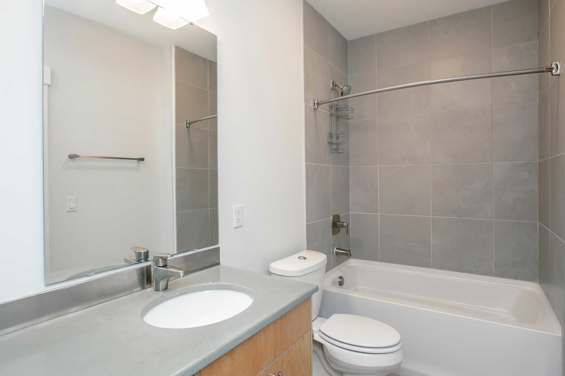 A modern bathroom featuring a bathtub with a curved shower rod, a toilet, and a single sink with a light-colored countertop. The walls are covered with large gray tiles, and there is a mirror above the sink reflecting the space. The overall design is clean and contemporary.