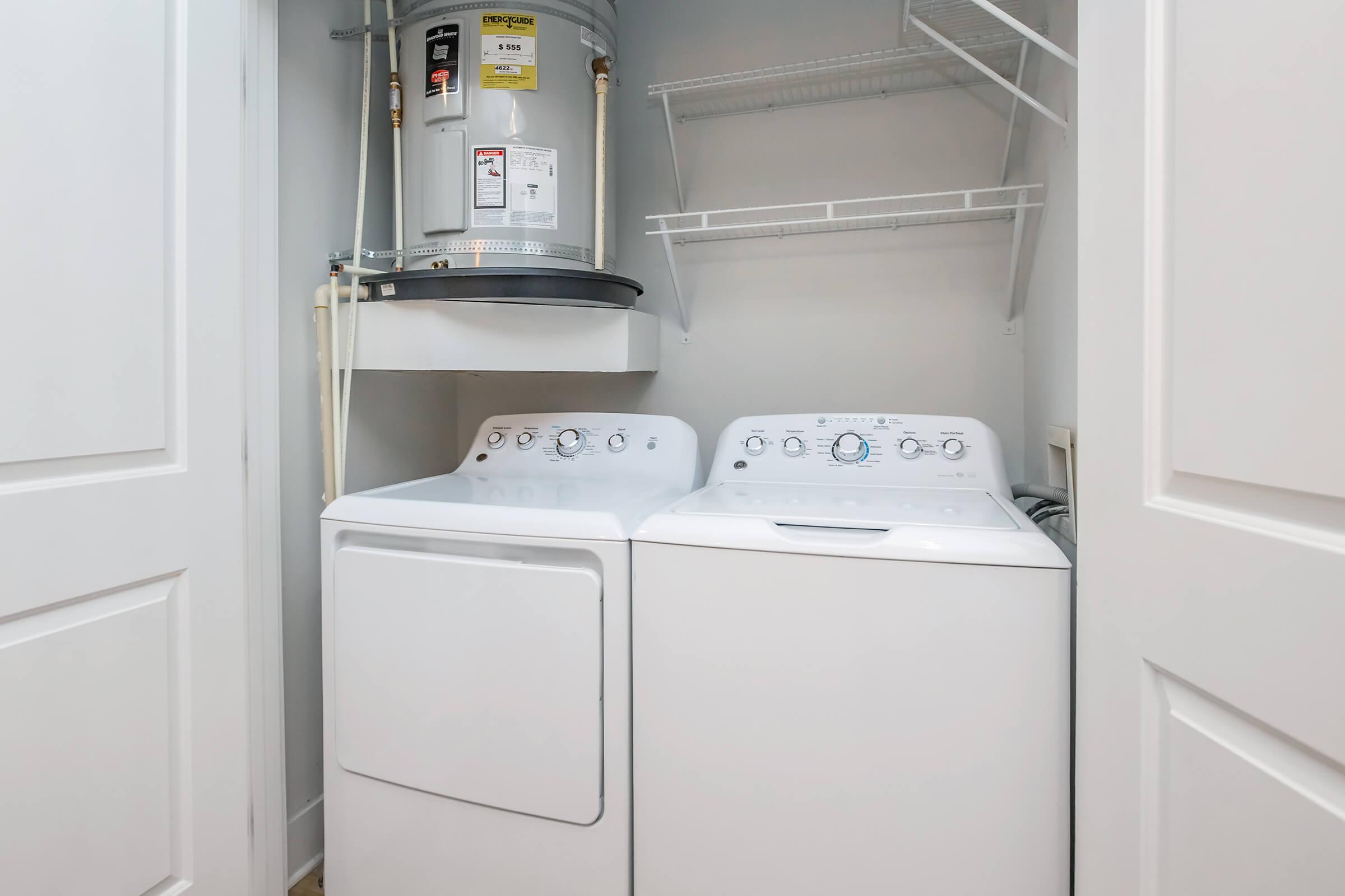 A clean laundry room featuring a stacked white washer and dryer side by side, with a water heater mounted above. There are white shelving units for storage, and the walls are painted in a light color, creating a tidy and organized space.