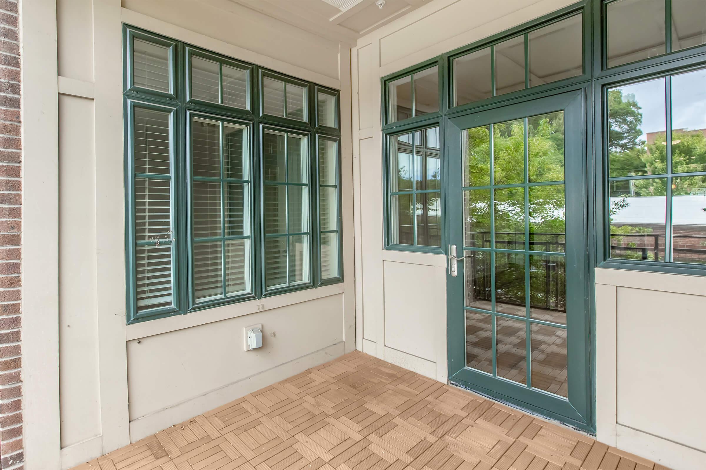 A cozy porch with large, green-framed windows, featuring a wooden floor made of interlocking tiles. The area is well-lit with natural light coming through the glass panes, and there's greenery visible outside, creating a serene and inviting atmosphere.