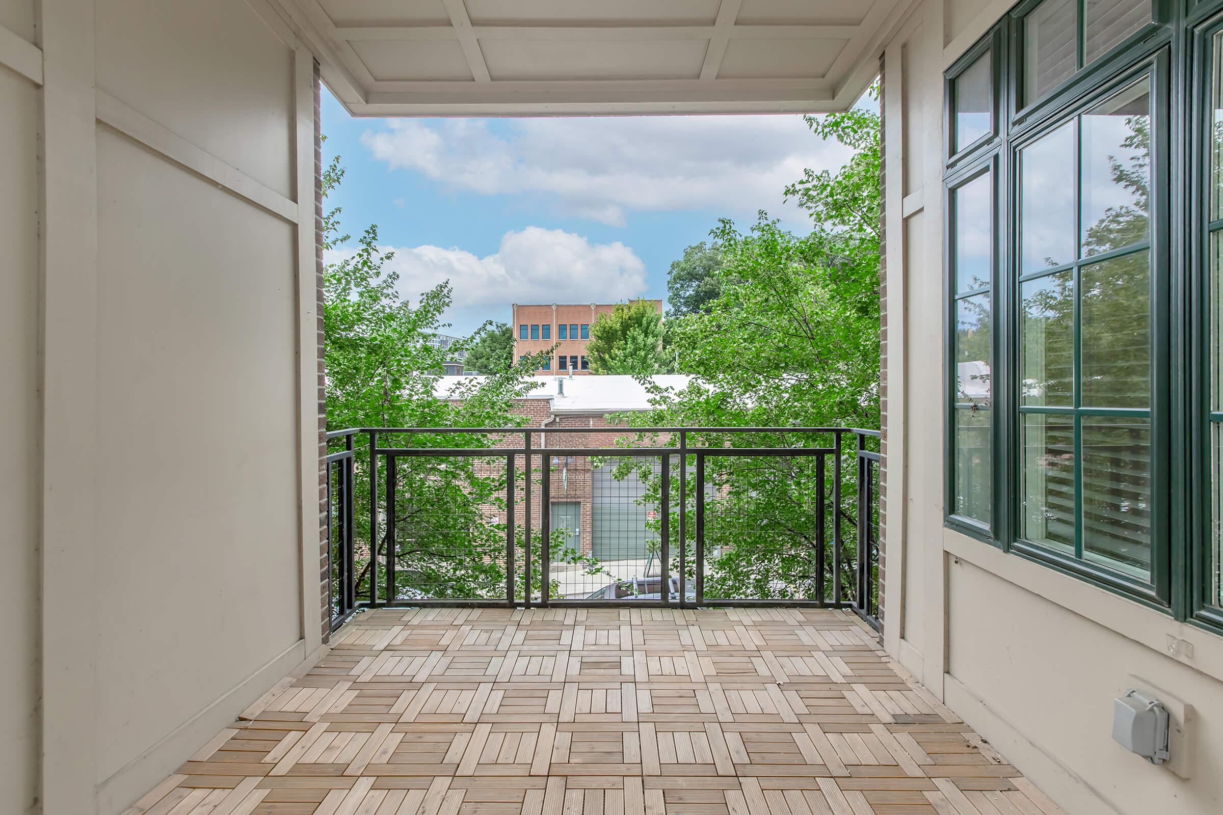 A view from a balcony featuring a wooden tile floor, surrounded by greenery. In the background, a building is partially visible under a cloudy sky. The balcony railing is black metal, and large windows on the sides provide additional natural light.