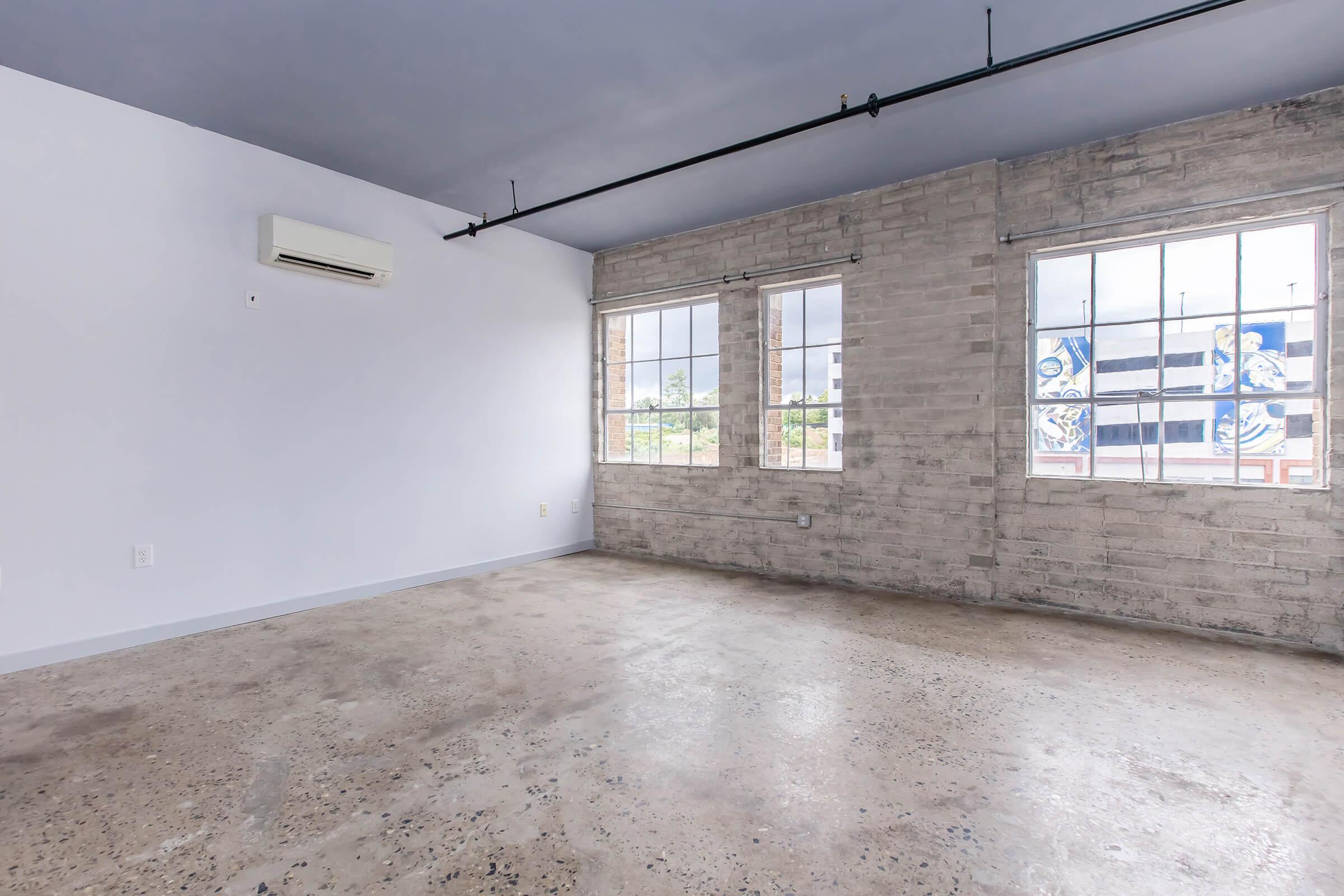 An empty industrial-style room featuring concrete flooring, exposed brick walls, and large windows that let in natural light. The ceiling is painted blue, and a wall-mounted air conditioning unit is visible. The overall aesthetic is minimalist and modern, ideal for various uses like an office or creative space.