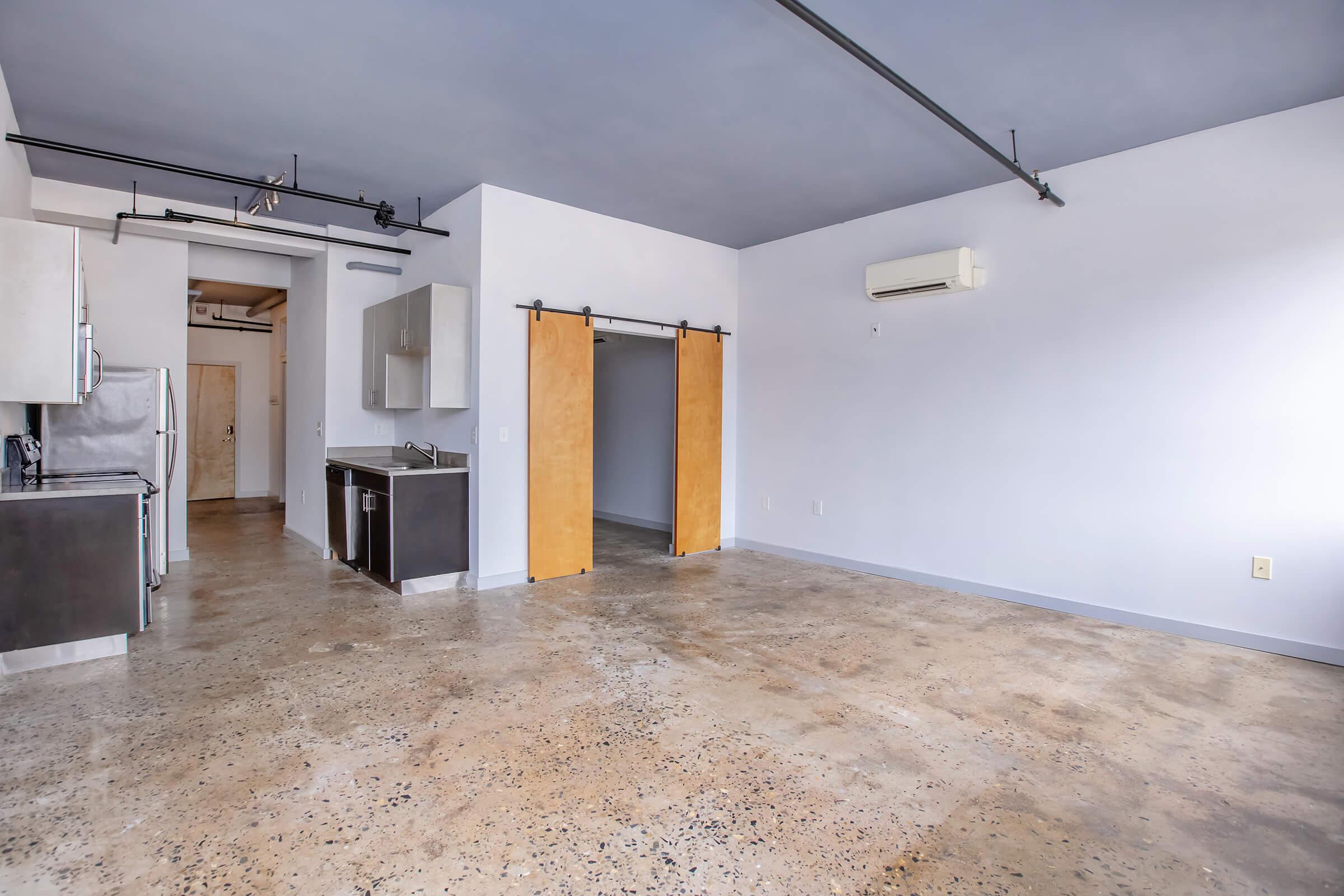 A spacious, modern apartment with polished concrete floors and high ceilings. The room features a kitchenette with stainless steel appliances, a sliding barn door, and minimalist decor. The walls are painted white, and the ceiling has a light blue color, providing a bright and airy atmosphere.