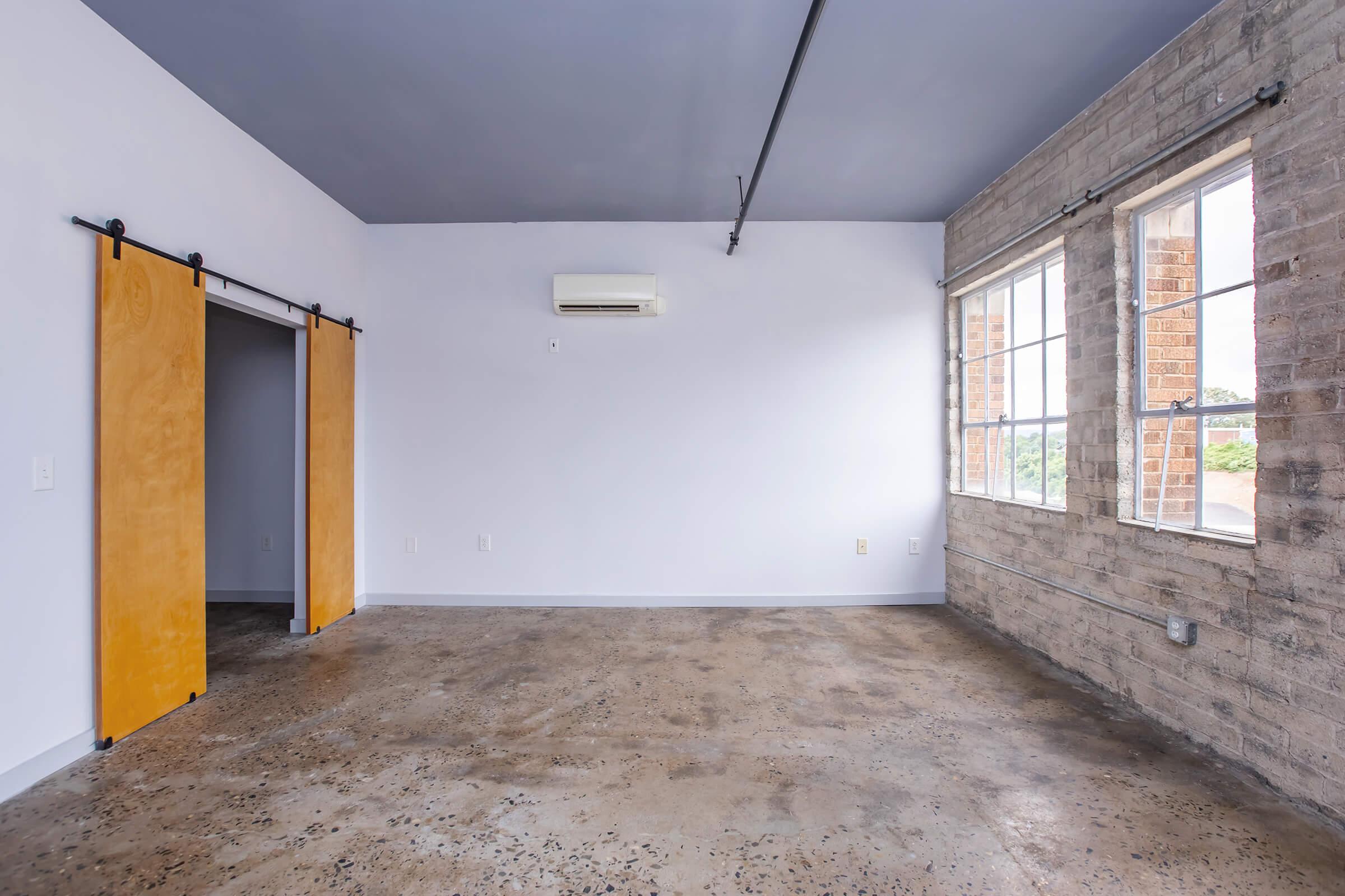 A spacious, minimalistic room featuring exposed brick walls and polished concrete floors. Two large windows allow natural light to enter, and there are sliding wooden barn doors leading to a closet. The ceiling is painted gray, and an air conditioning unit is mounted on the wall, enhancing the modern aesthetic.