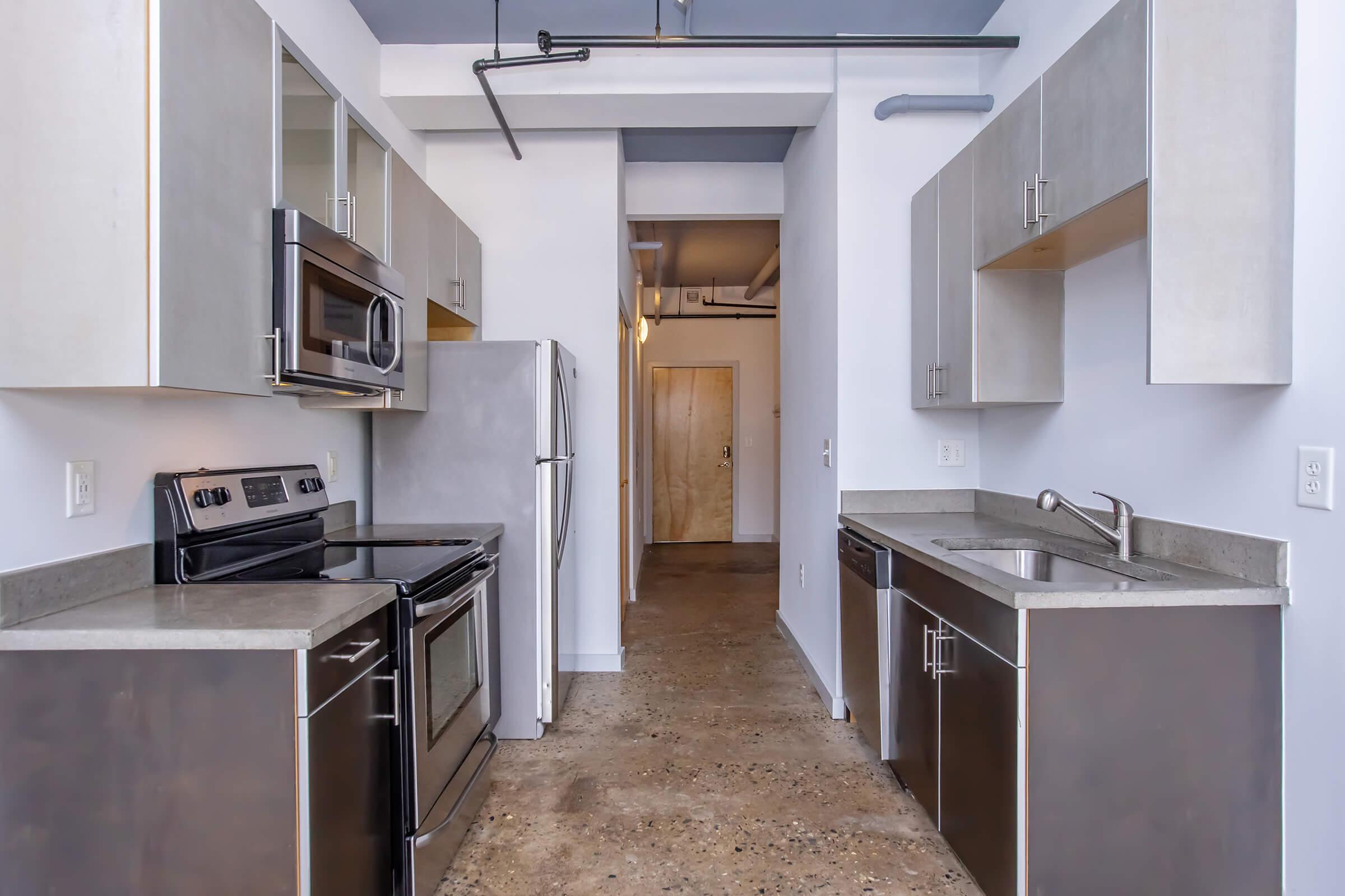 Modern kitchen featuring stainless steel appliances including a microwave, oven, and refrigerator. The cabinetry is a sleek combination of light and dark finishes. A concrete countertop contrasts with the cabinets. The space is designed with an open layout, leading to a hallway with a door at the end.