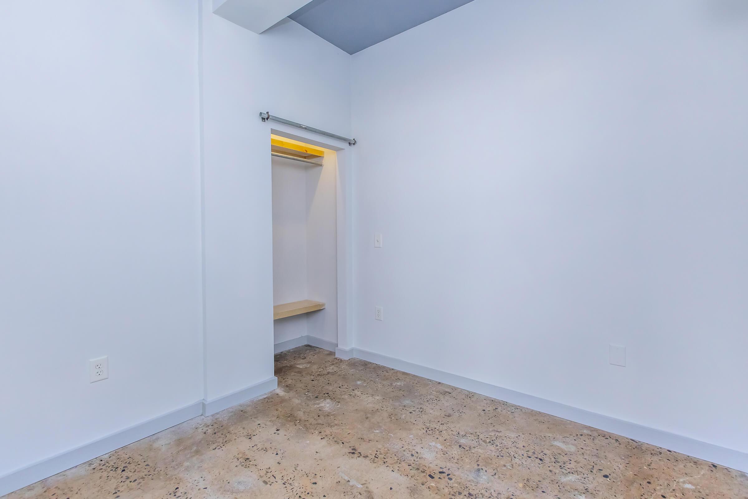 Empty room with light blue walls and a concrete floor. There is a small open closet with a wooden shelf, but no other furniture or decorations present. Natural light enters from a source above, creating a bright and airy atmosphere.