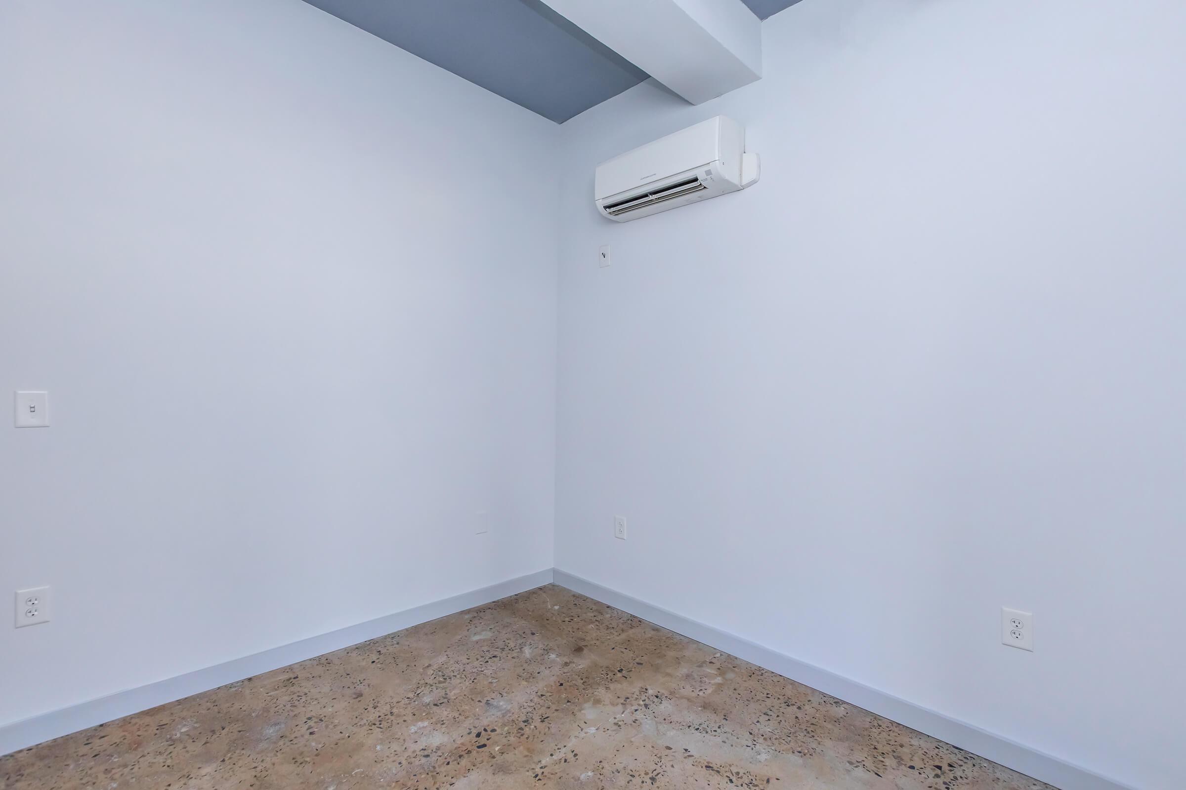 A well-lit, empty room with light blue walls and a concrete floor. An air conditioning unit is mounted on the wall, and there are electrical outlets on the wall. The space appears clean and minimalistic, suitable for various uses.