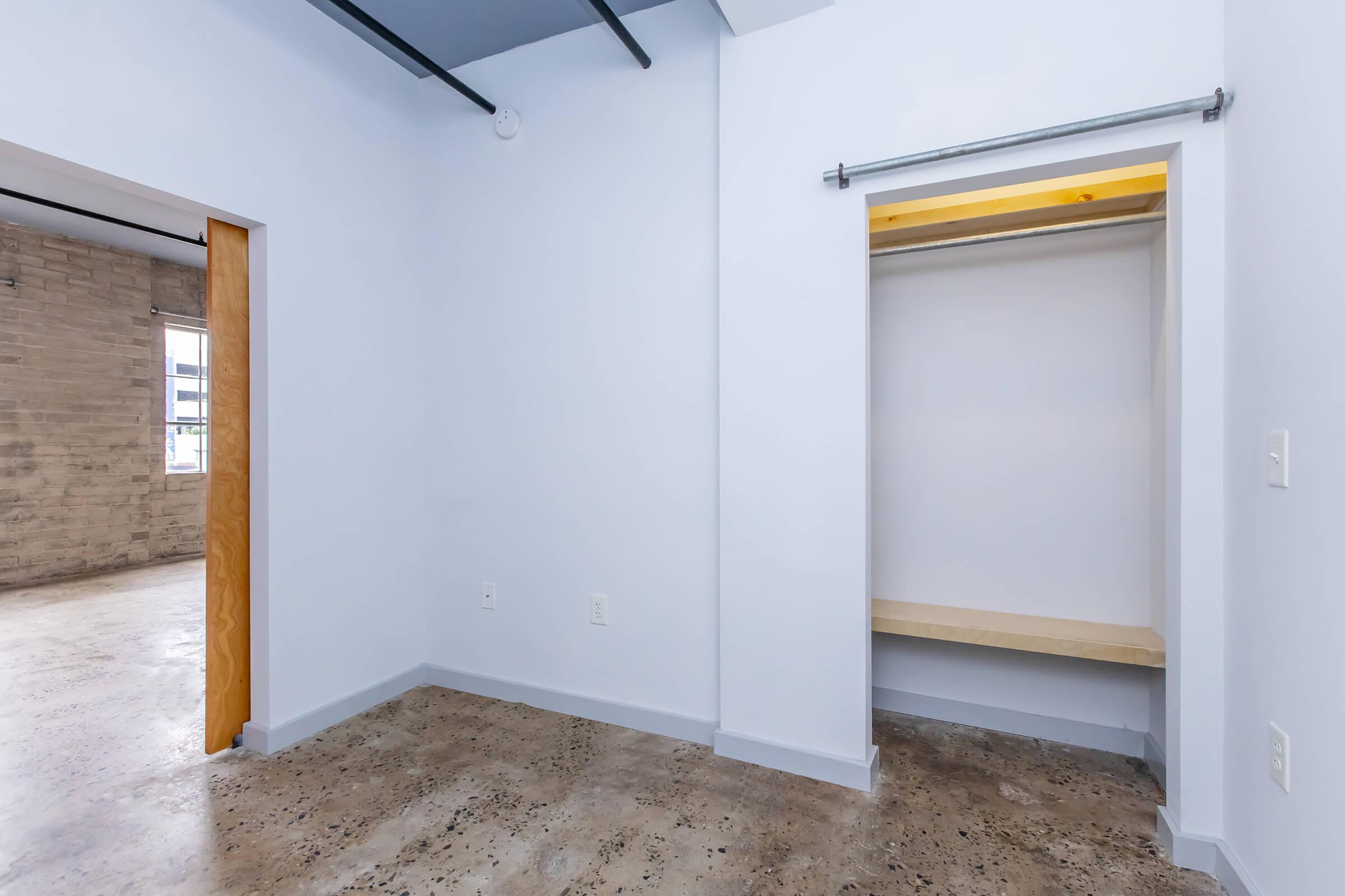 A view of a small room featuring light blue walls and a concrete floor. On the left, there is a wooden door leading to another area, while on the right, an open closet with a wooden shelf is visible. The space has minimalist decor and abundant natural light.