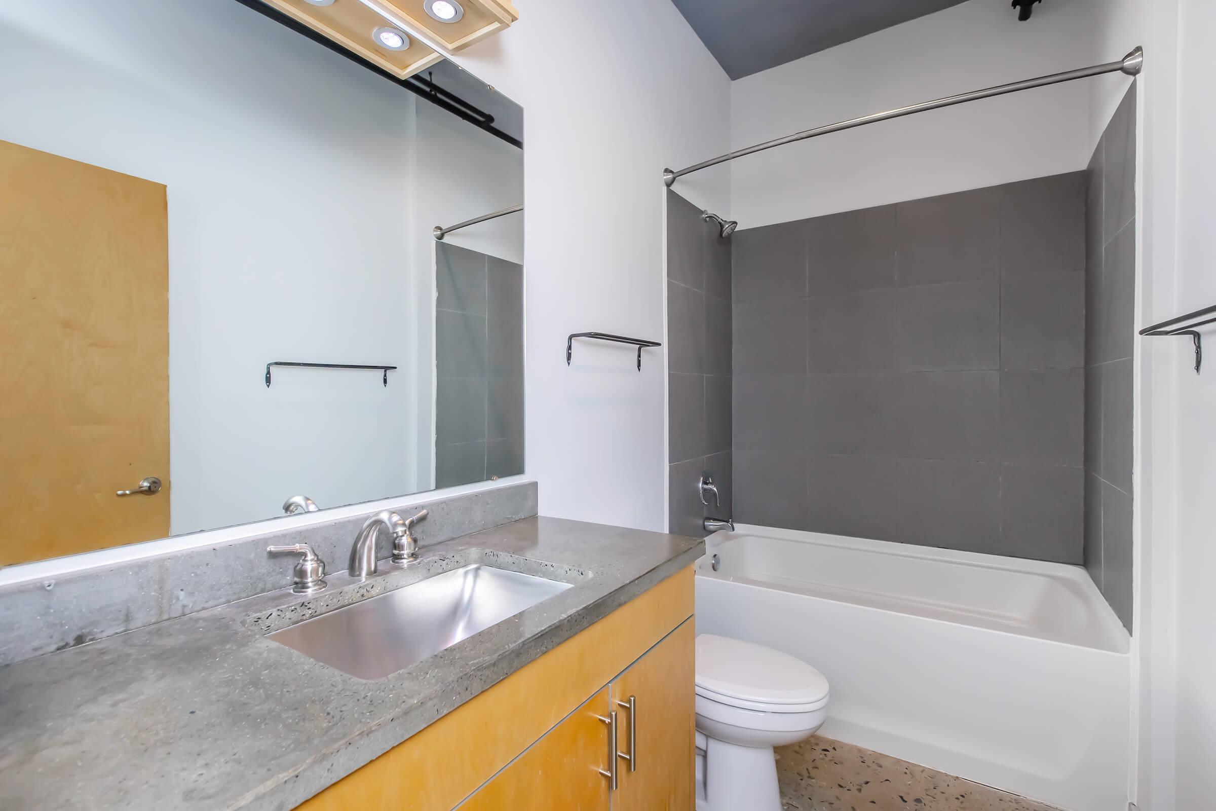 A modern bathroom featuring a sleek vanity with a concrete countertop, a mirror above, and a silver faucet. The shower area has gray tiles and a glass shower rod. A bathtub is visible alongside a white toilet, set against a light-colored wall and floor with a minimalist design.