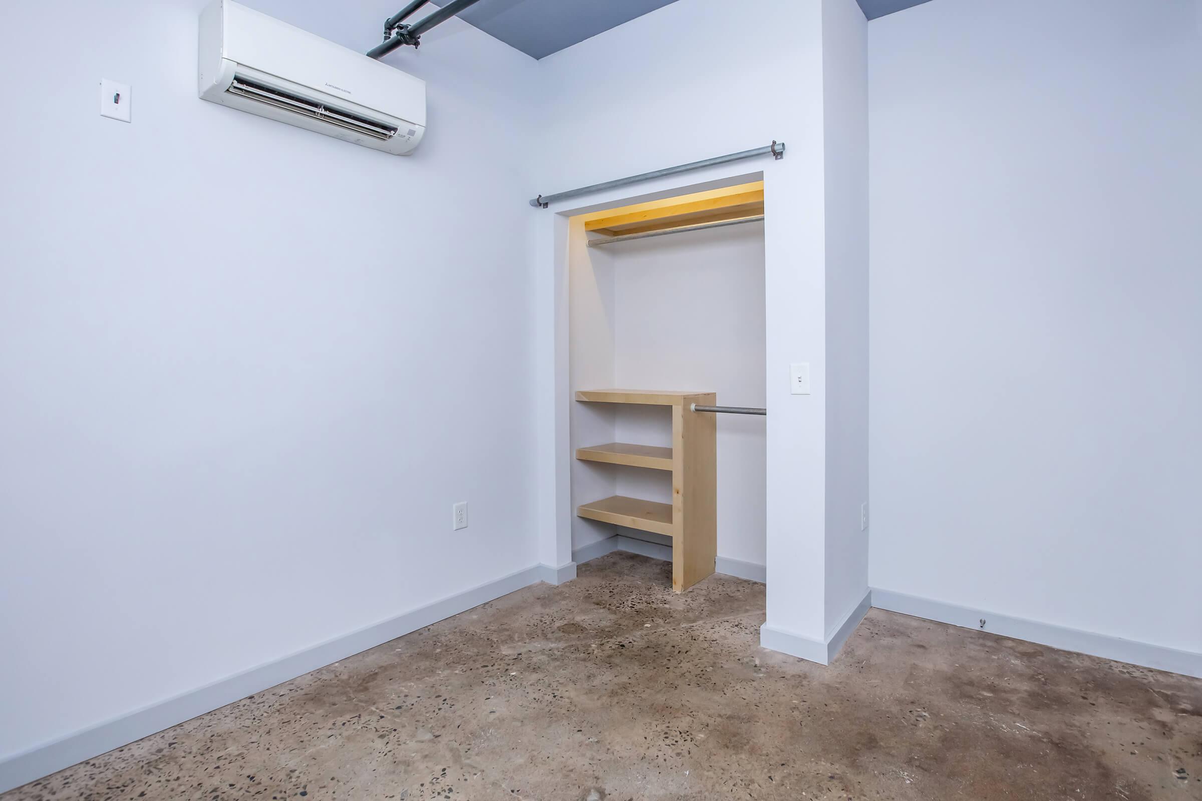 Empty closet with wooden shelves and a hanging rod, featuring a light wall color and a concrete floor. An air conditioning unit is mounted on the wall above.